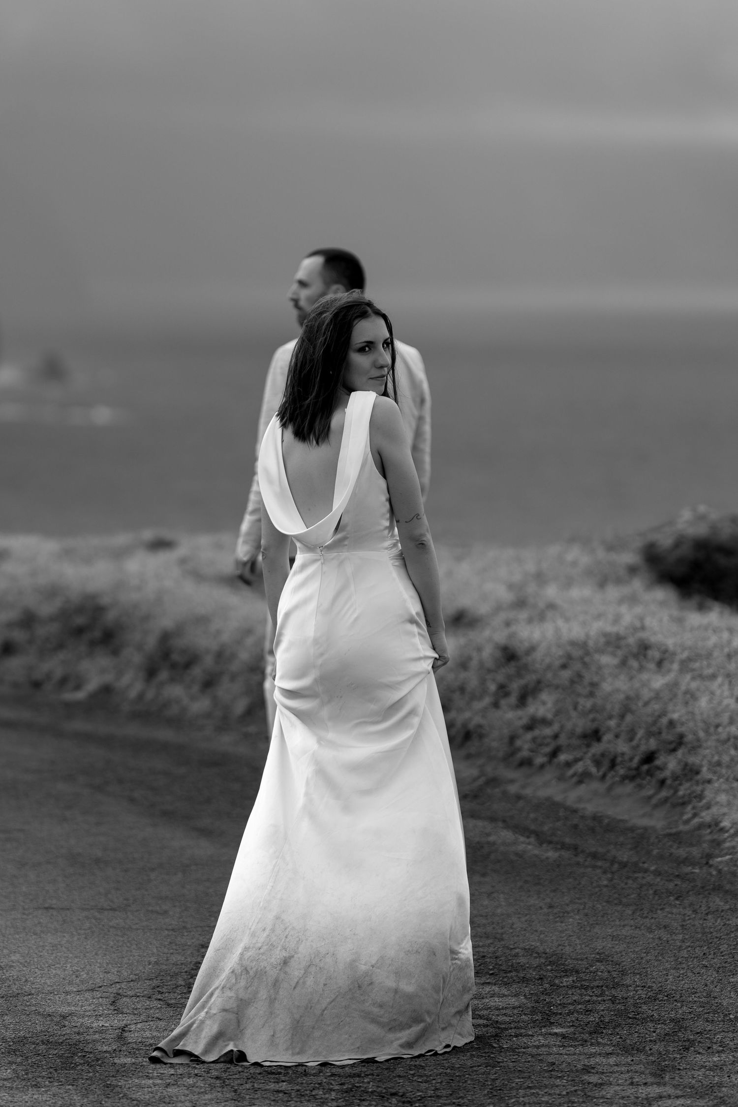 Black and white photo of bride in open-back wedding gown walking with groom along a coastal road in Madeira.
