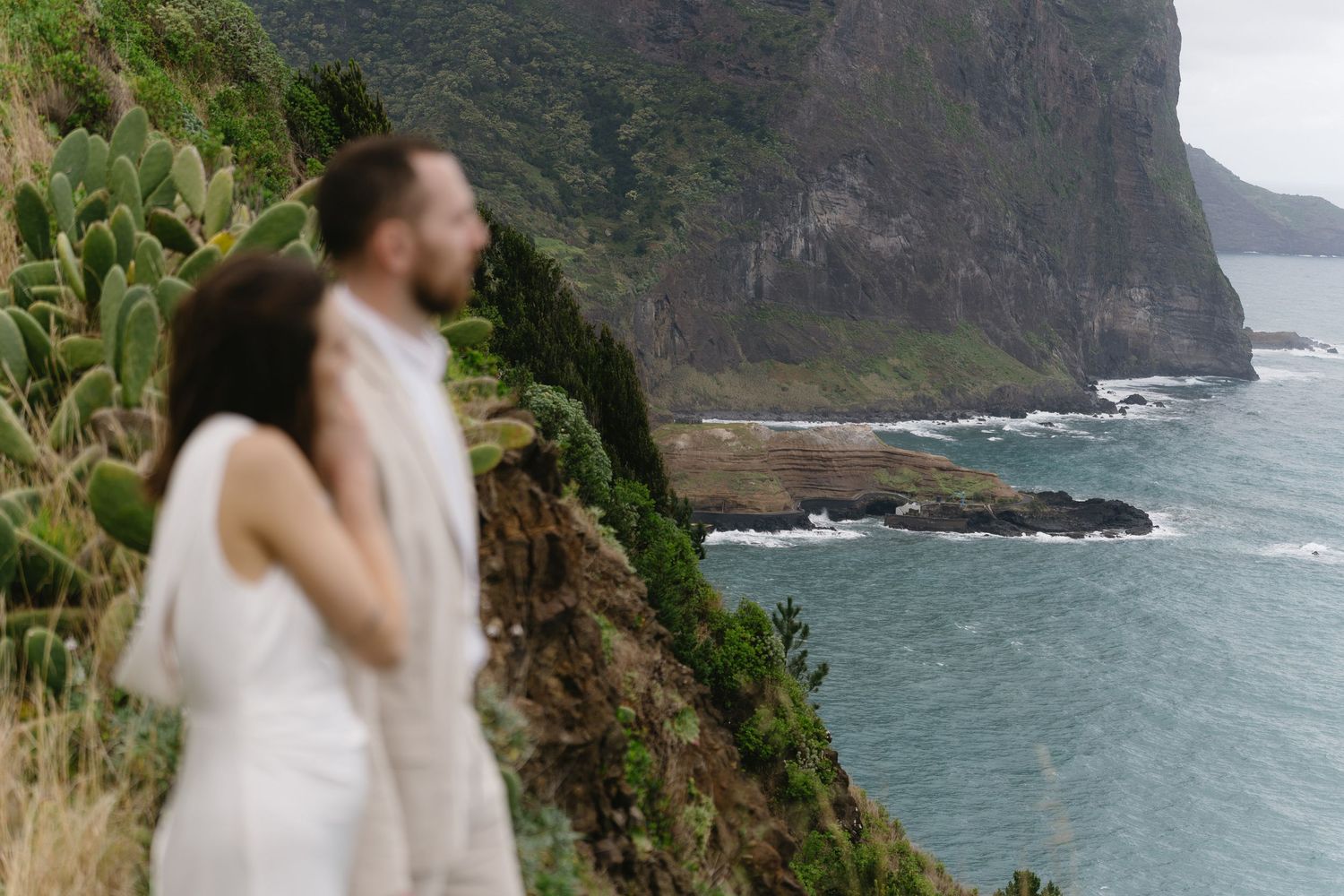 Couple embracing with dramatic green coastal cliffs and turquoise ocean behind them.