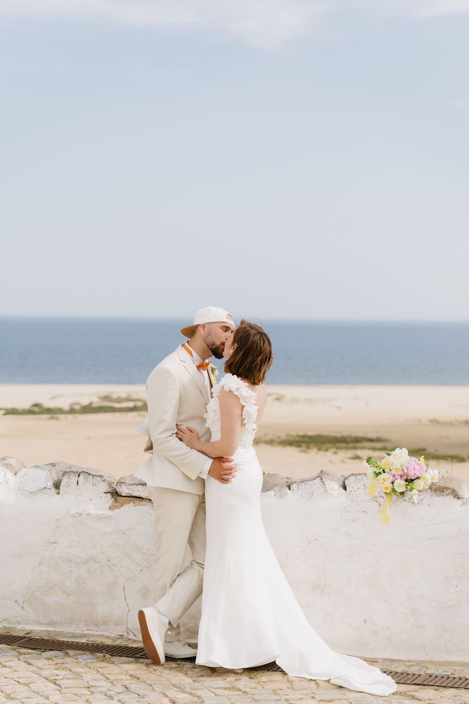 Newlyweds share a kiss near stone wall with ocean and sandy beach in background.