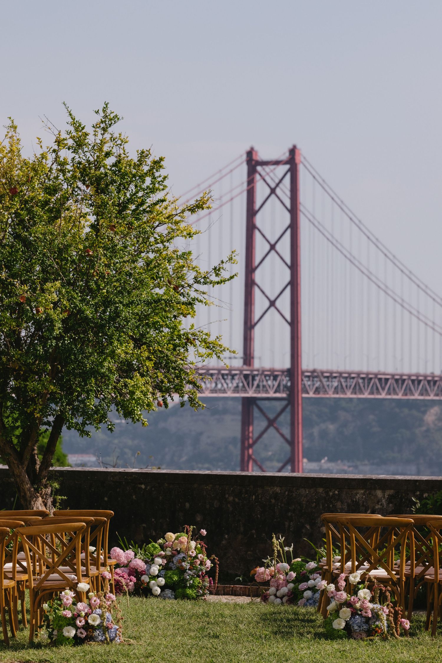 Outdoor wedding ceremony setup with wooden chairs and floral arrangements, overlooking the 25 de Abril Bridge in Lisbon.
