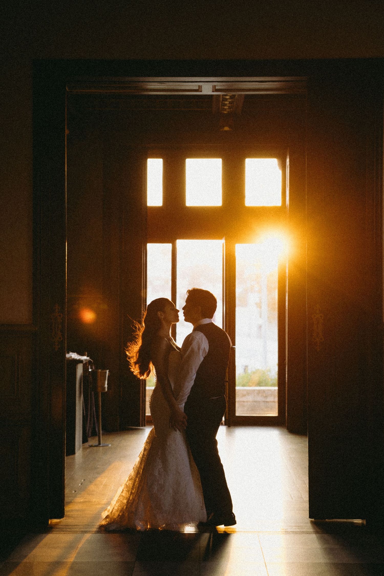 Silhouette of bride and groom sharing a romantic moment in golden sunset light streaming through elegant doorway.