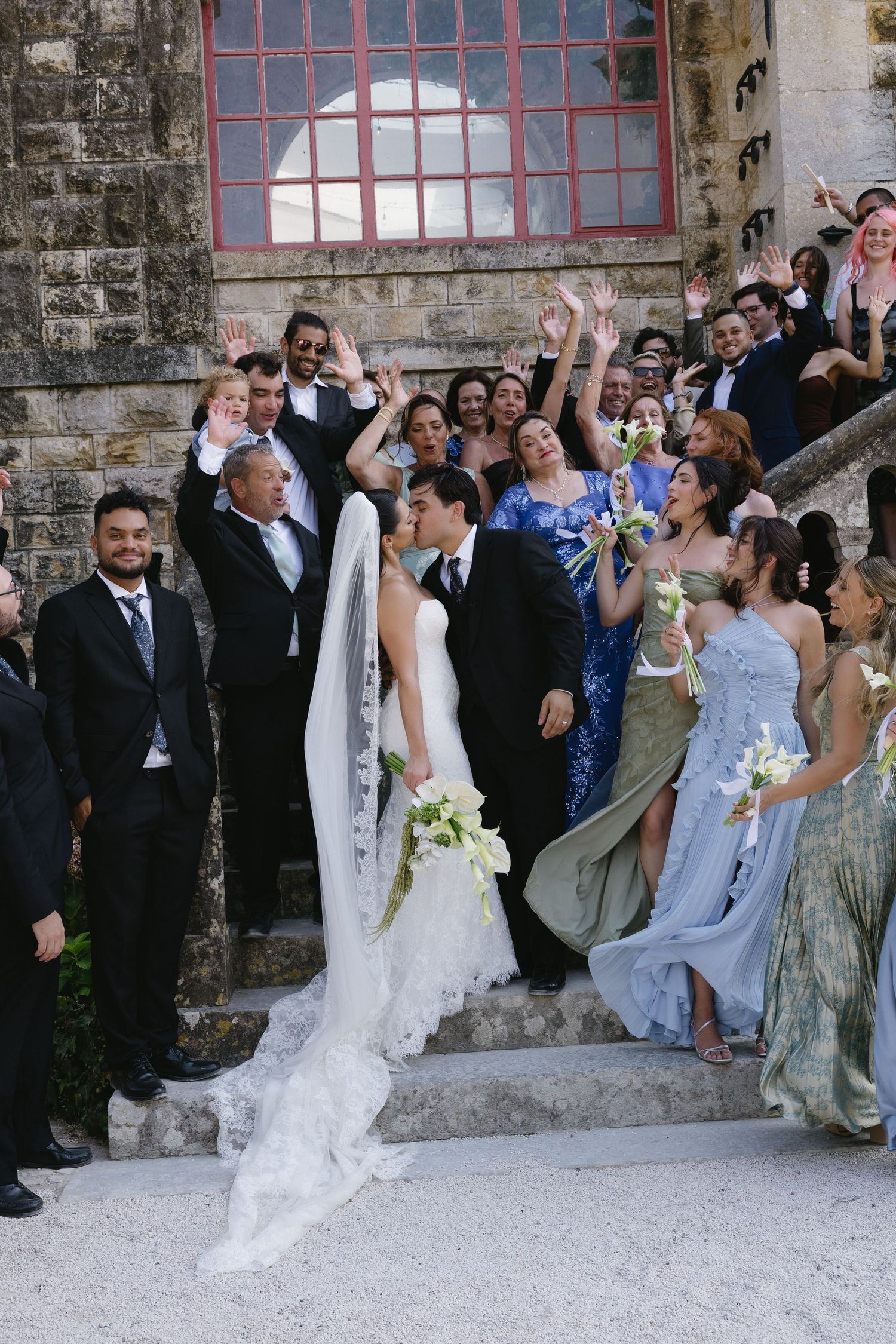 Bride and groom kiss on stone steps surrounded by cheering wedding party and guests at elegant venue.