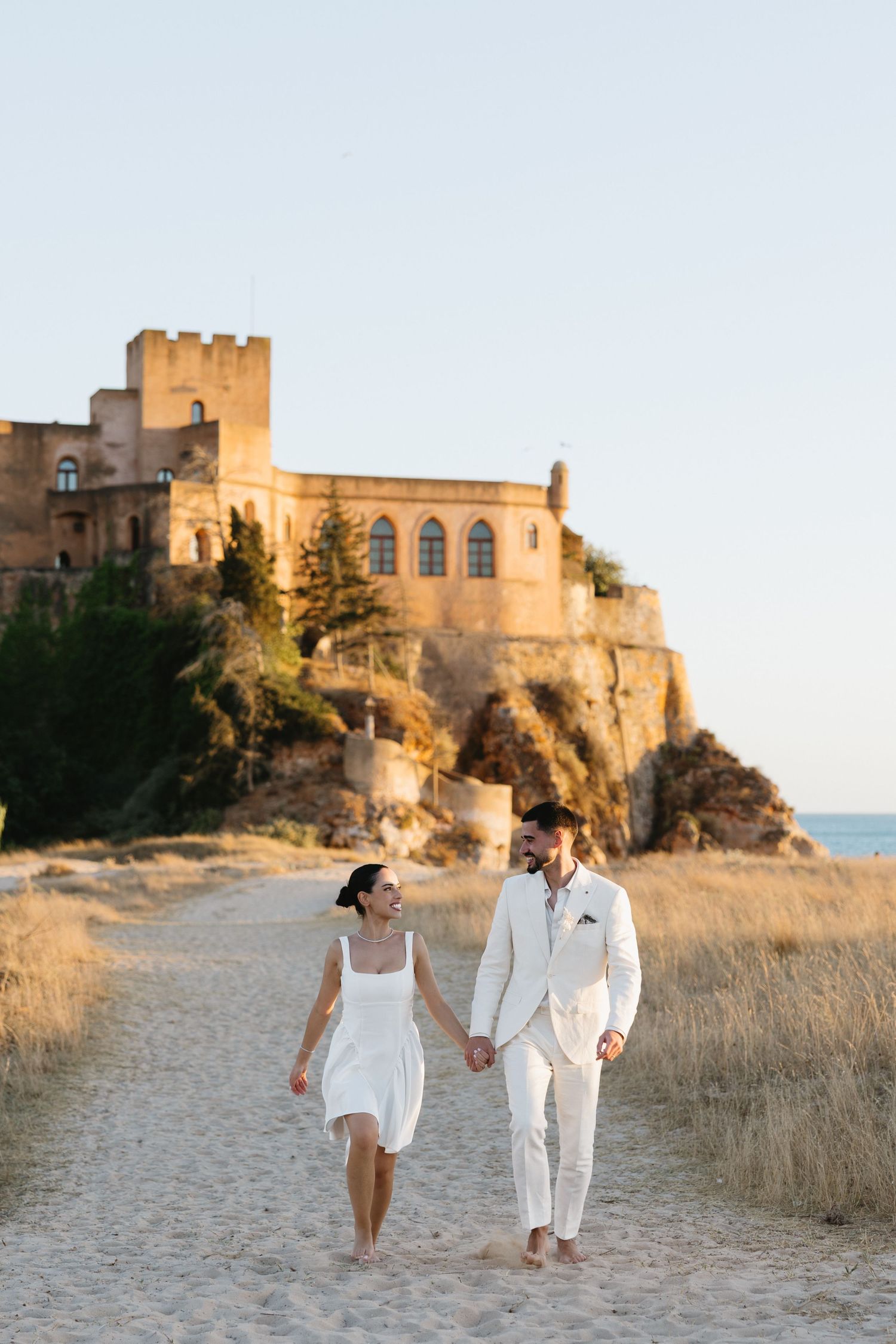 Bride and groom walking during their wedding at praia da angrinha with the castle of ferragudo in the background, Algarve