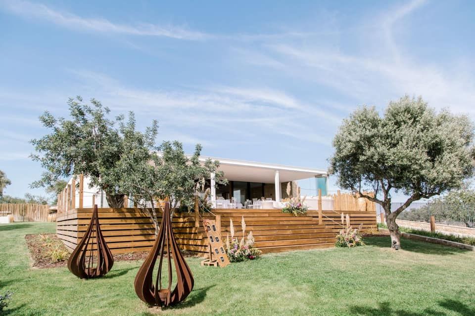 Contemporary wooden deck venue surrounded by lush green lawn, olive trees, and decorative metal sculptures.