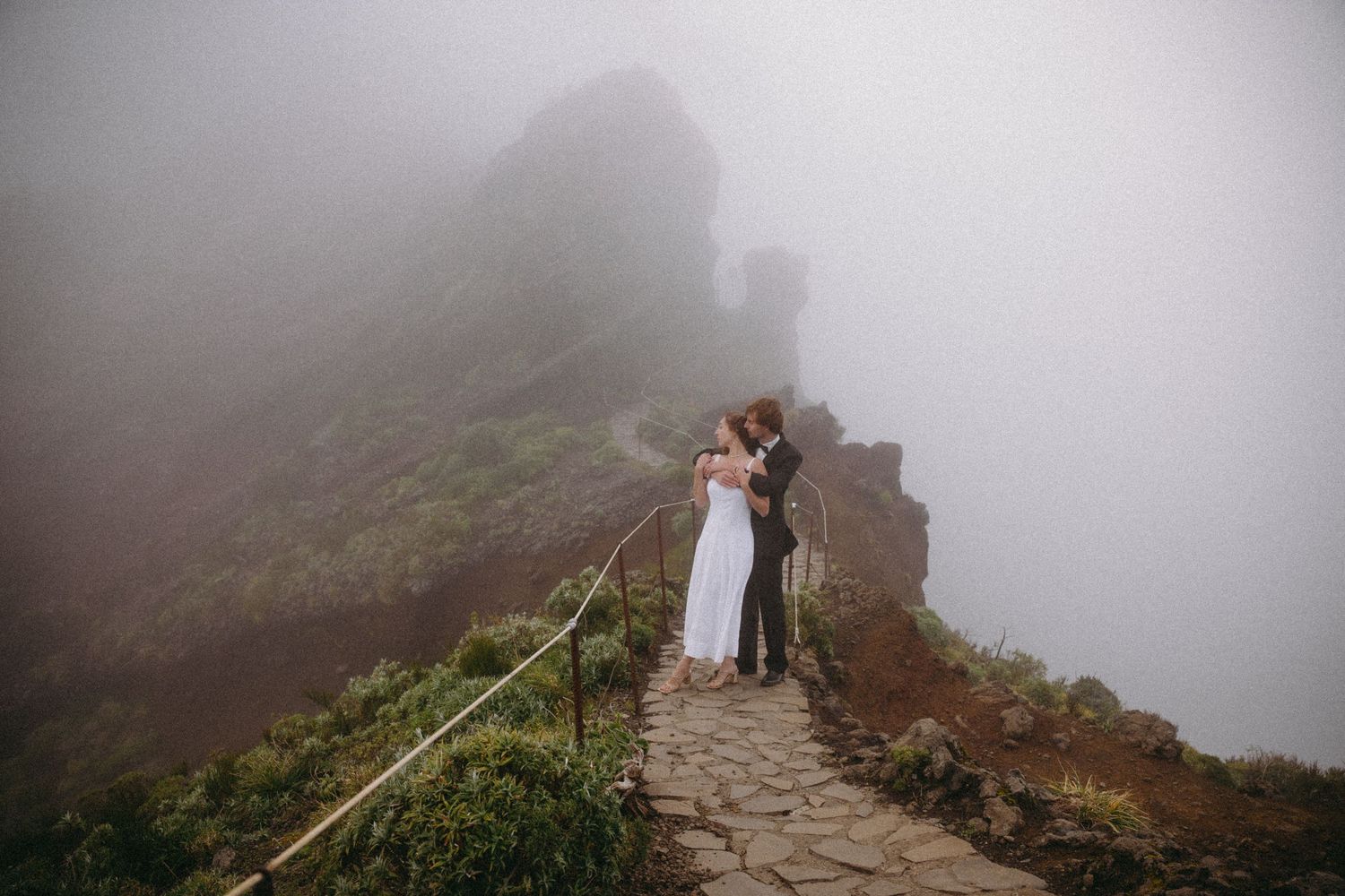 Wedding couple embracing on a dramatic misty mountain ridge path with rocky cliffs.