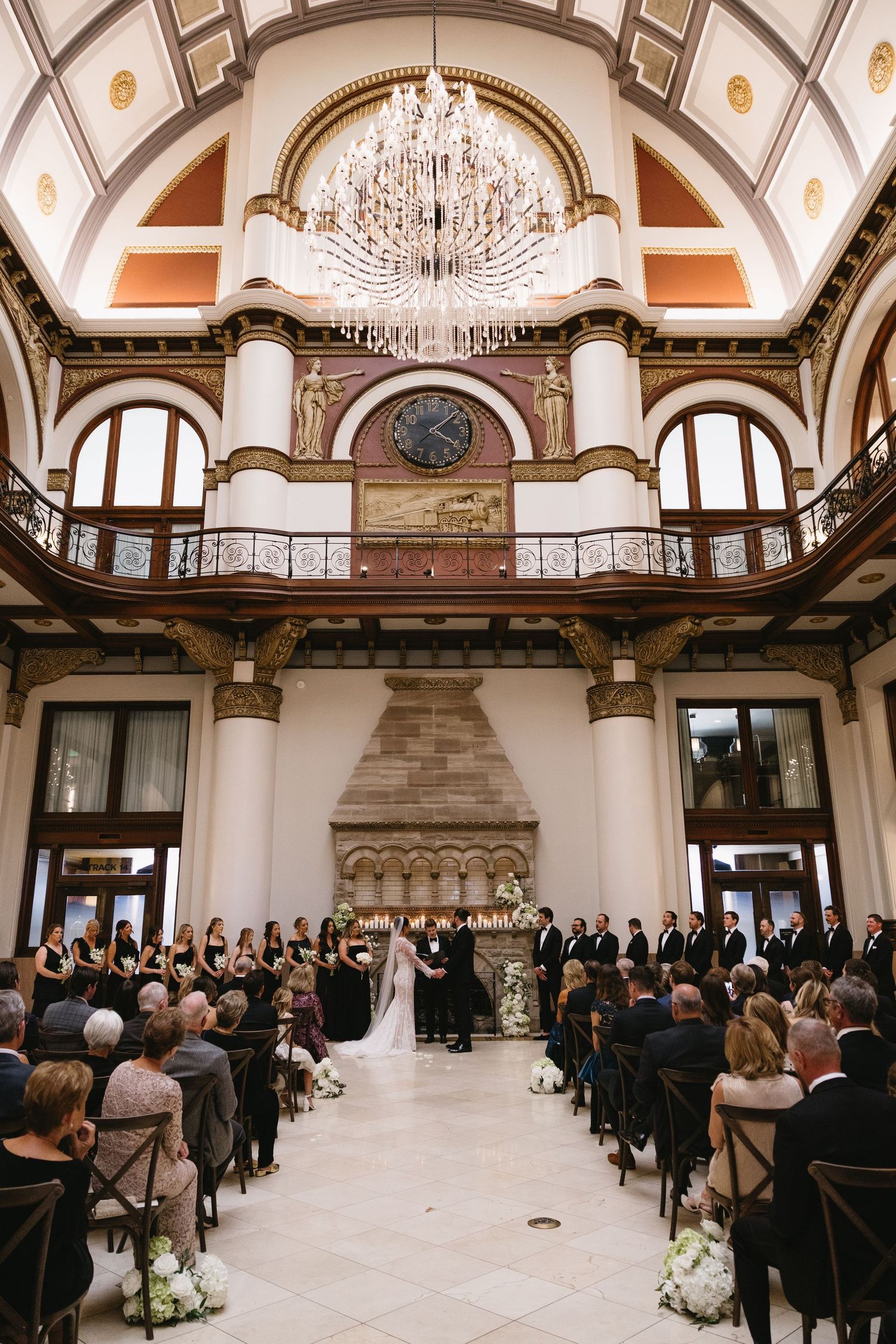 Elegant wedding ceremony in grand ballroom with chandelier, fireplace, floral arrangements, and guests seated in wooden chairs.