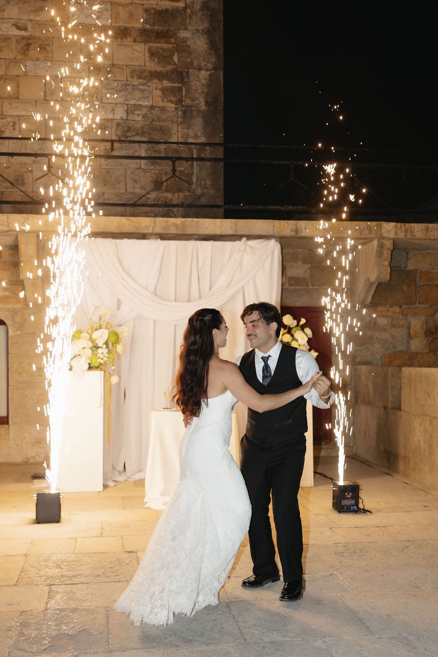 Bride and groom share first dance surrounded by sparkling fountain fireworks at elegant stone venue wedding reception.