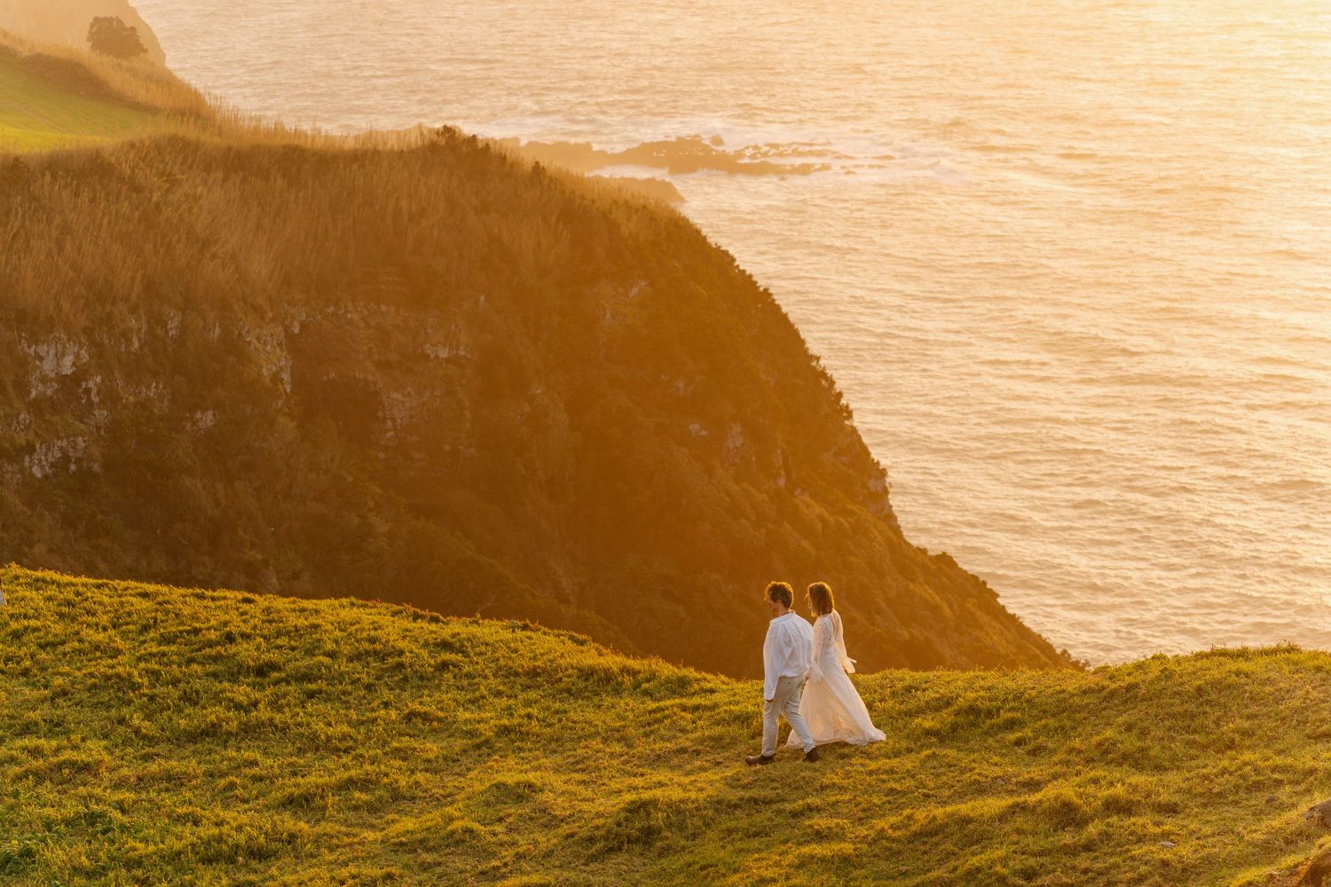 Bride and groom walking hand in hand along a dramatic coastal cliff at golden sunset overlooking the ocean.