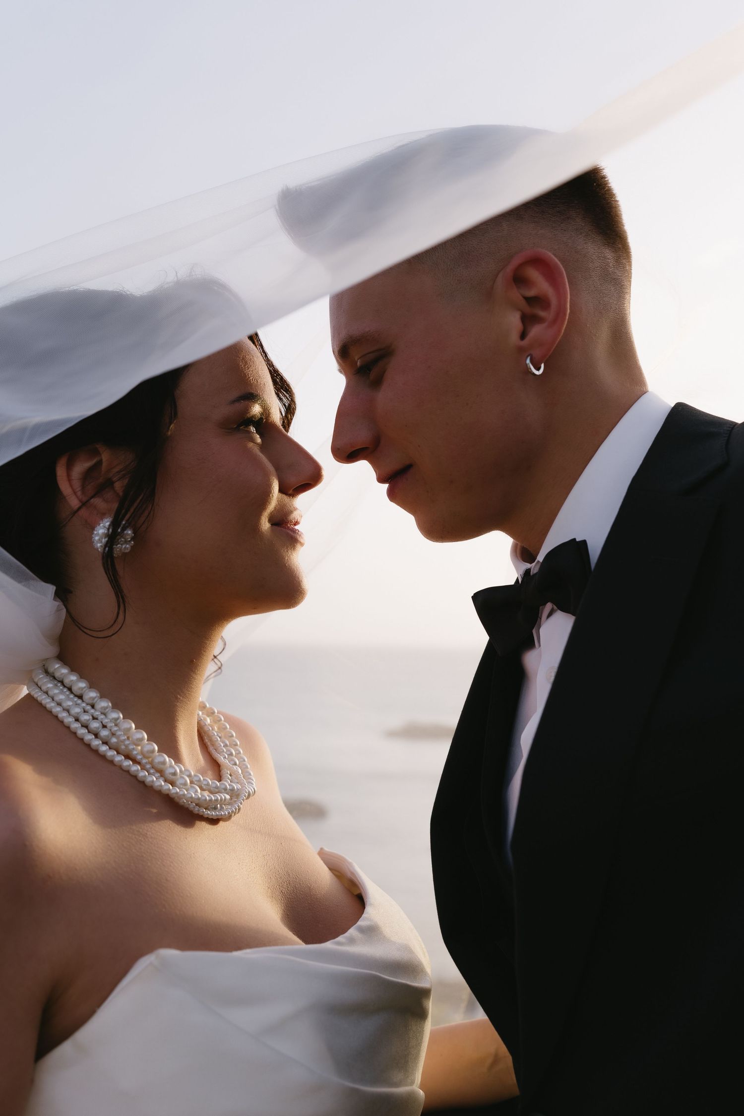 Intimate close-up of bride and groom almost kissing under a veil, bride wearing pearl necklace by the sea.