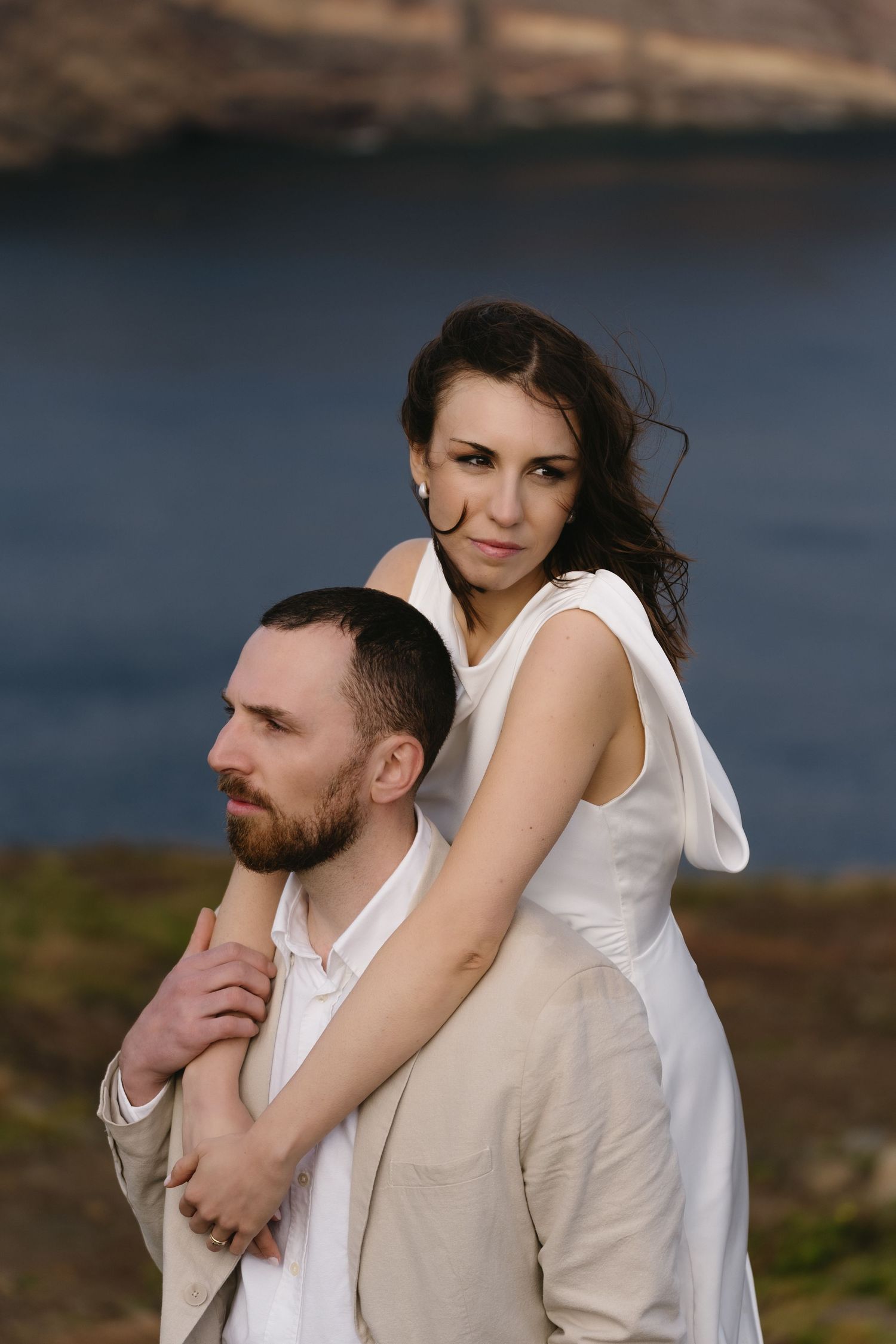 Bride embracing groom from behind near a serene blue lake backdrop.