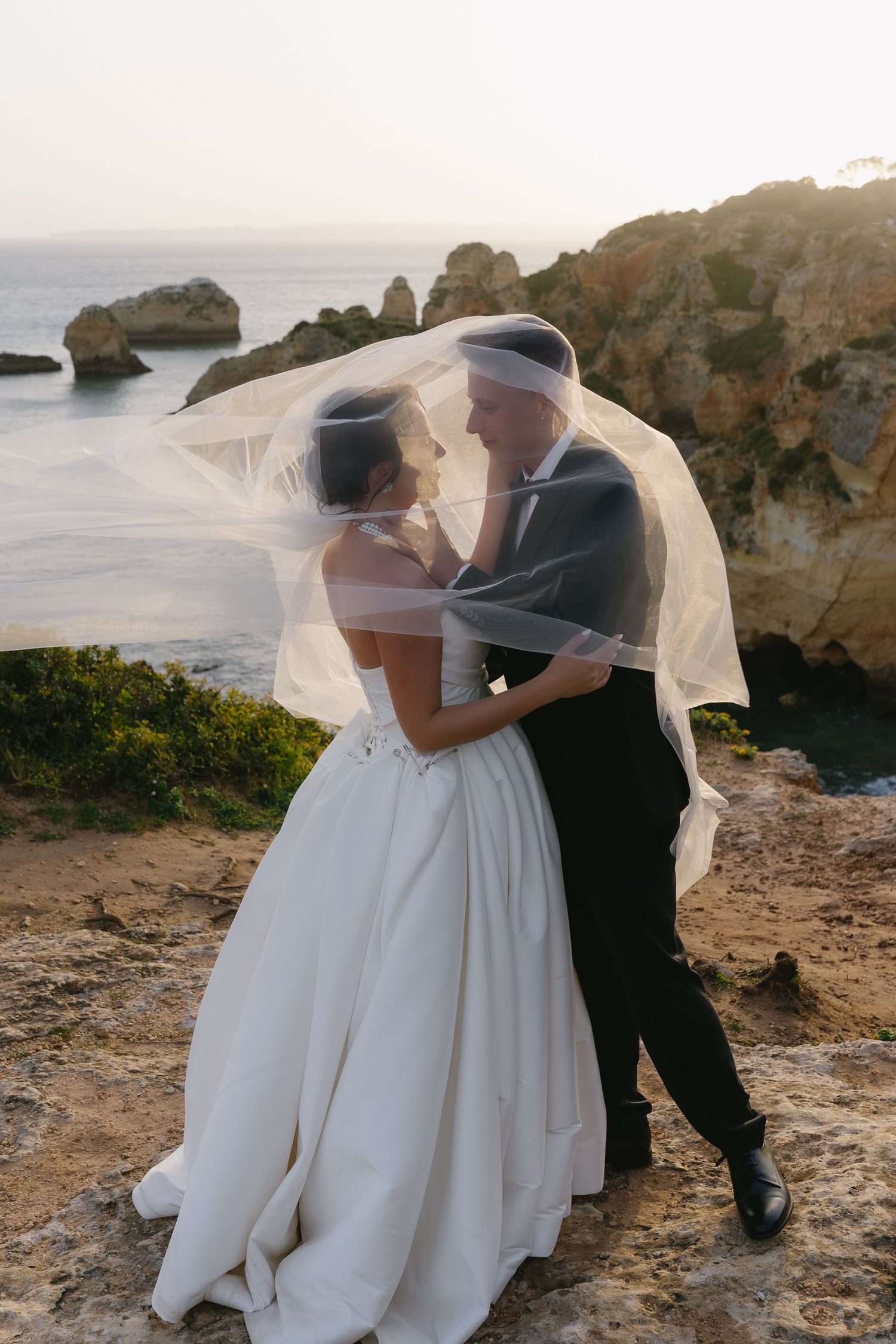 Bride and groom share a romantic moment under a flowing veil on a rocky coastal cliff at sunset.