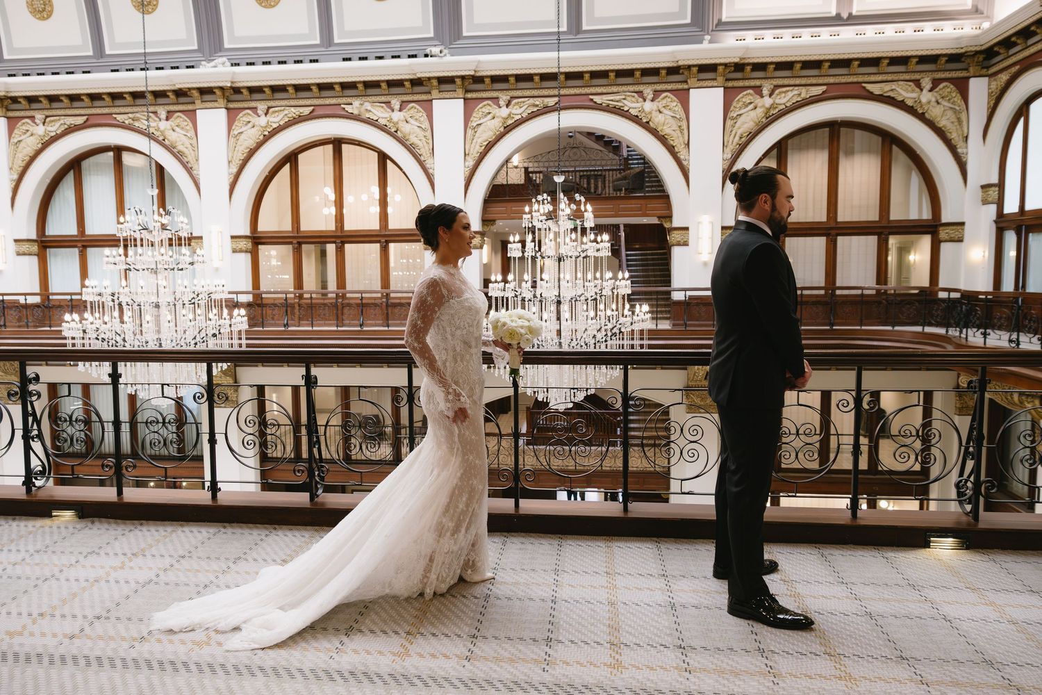 Bride and groom share a romantic first look moment in an elegant ballroom with chandeliers and ornate balcony.