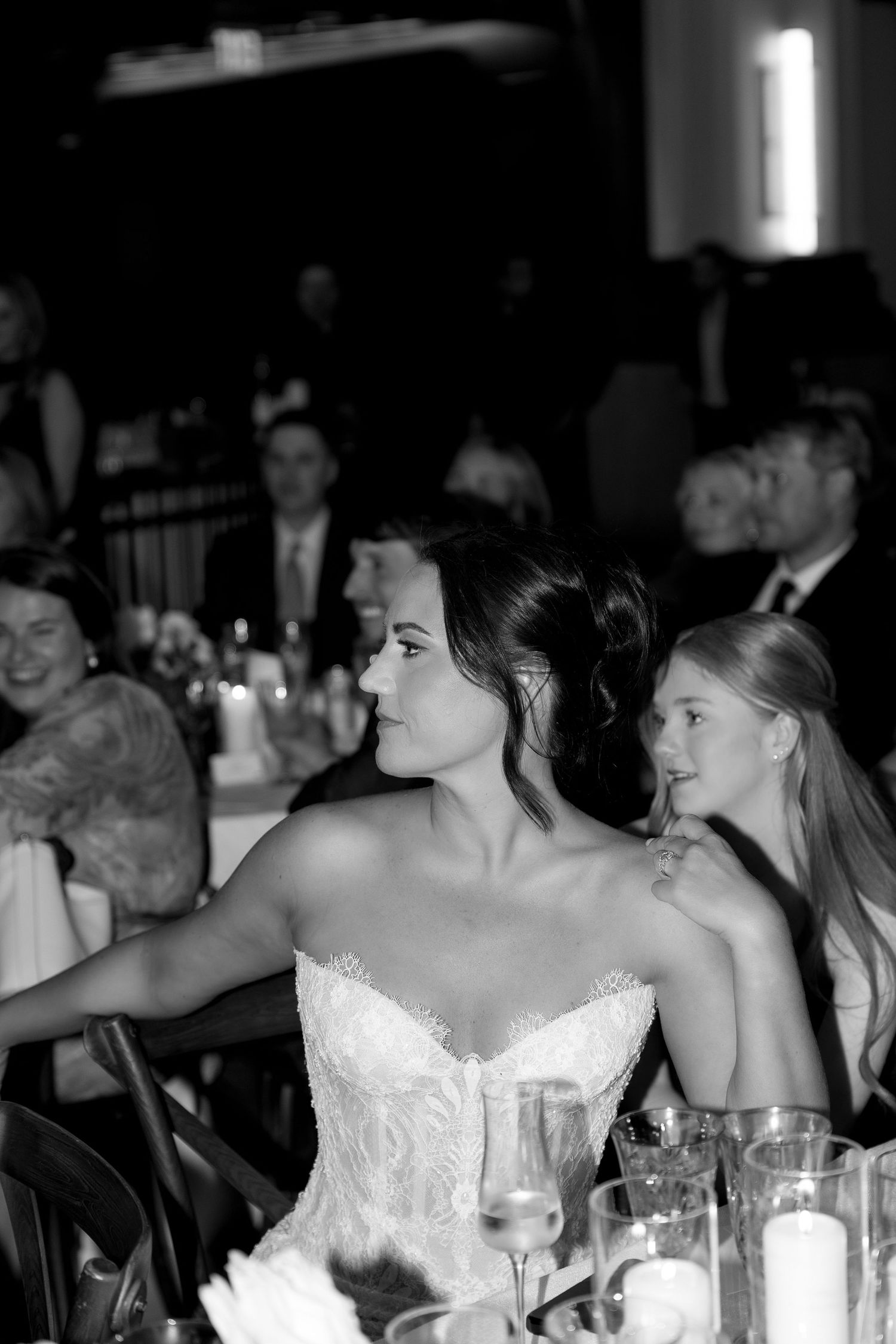 Bride in strapless lace gown seated at wedding reception table, black and white photo with guests in background.