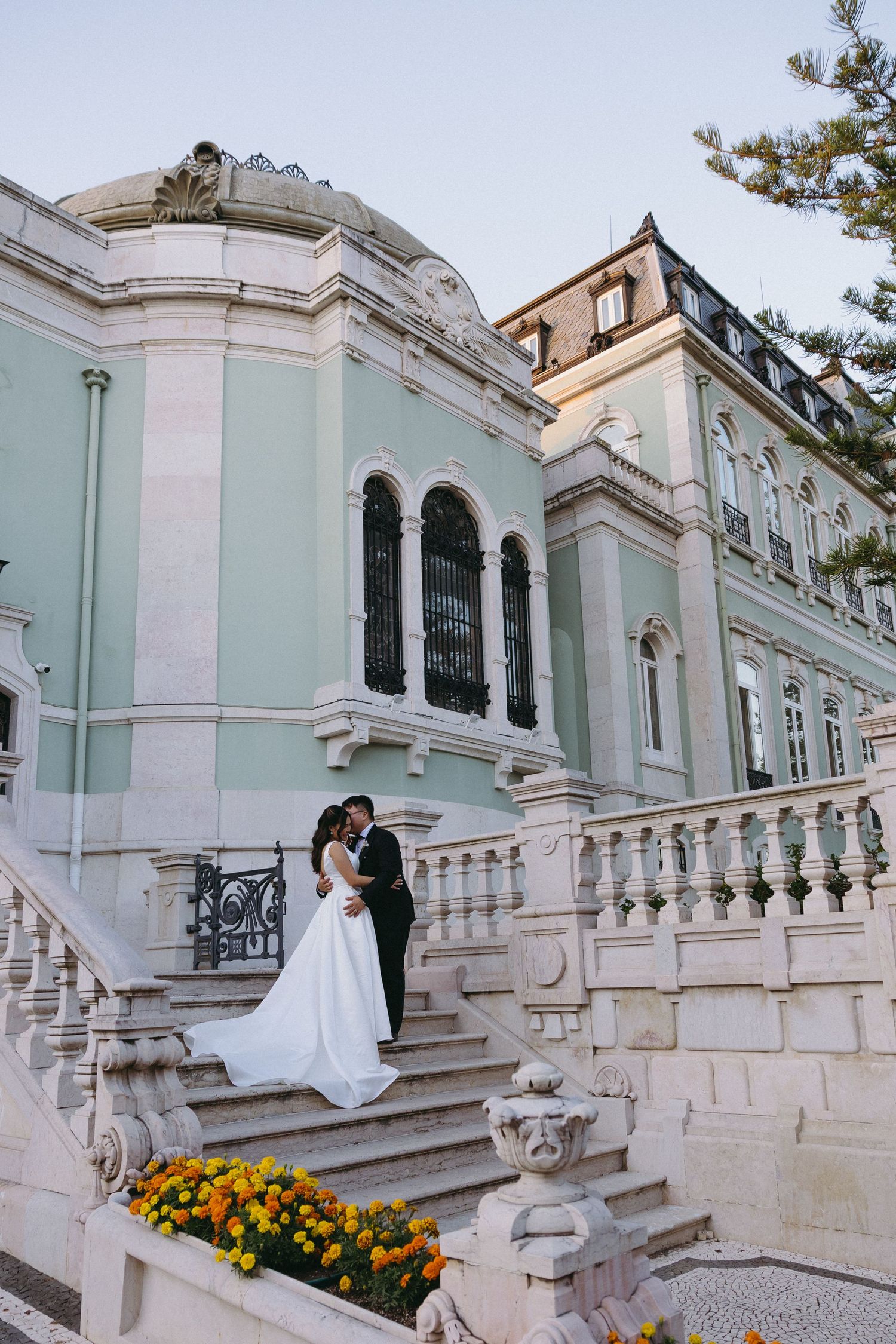 Bride and groom embrace on grand stone steps of elegant mint green European palace with ornate balustrade.