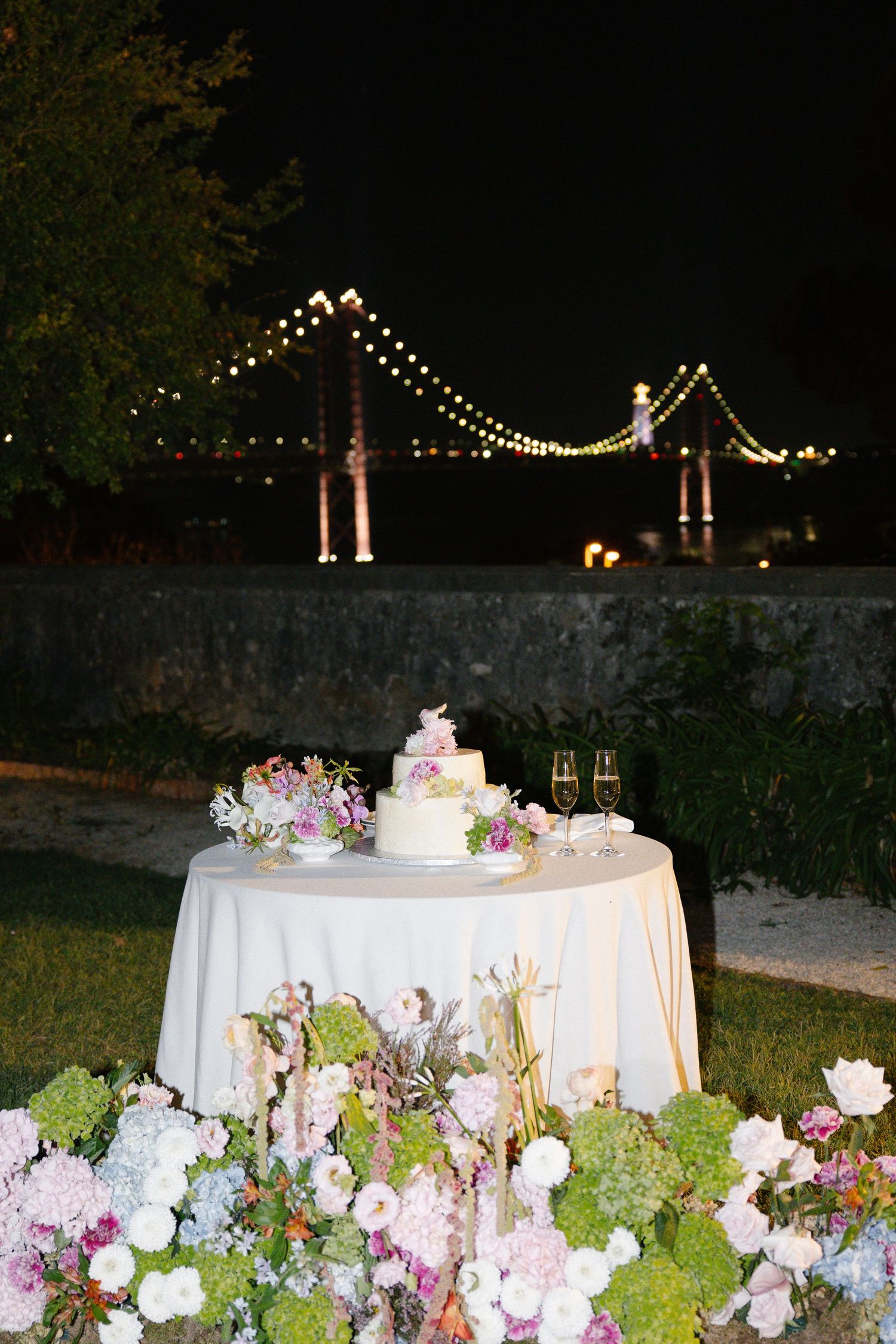 Elegant wedding cake table adorned with colorful flowers, champagne flutes, and a lit bridge backdrop at night.