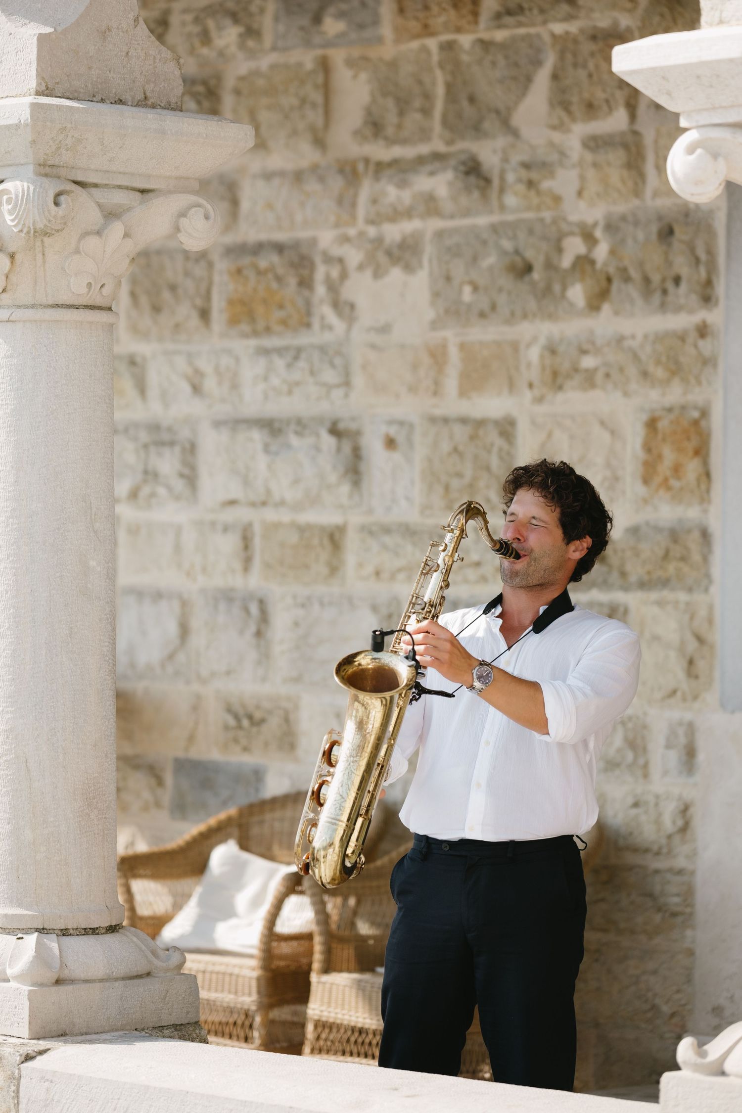 Man in white shirt playing saxophone outdoors against stone wall with elegant columns.