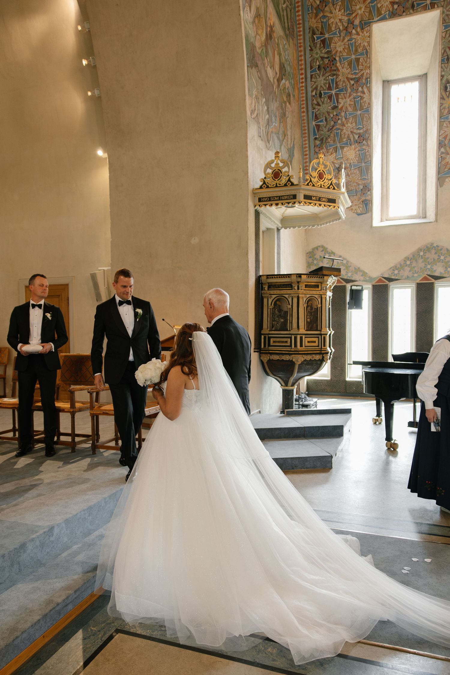 Bride in flowing white gown walks down church aisle with escort toward groom during elegant wedding ceremony.