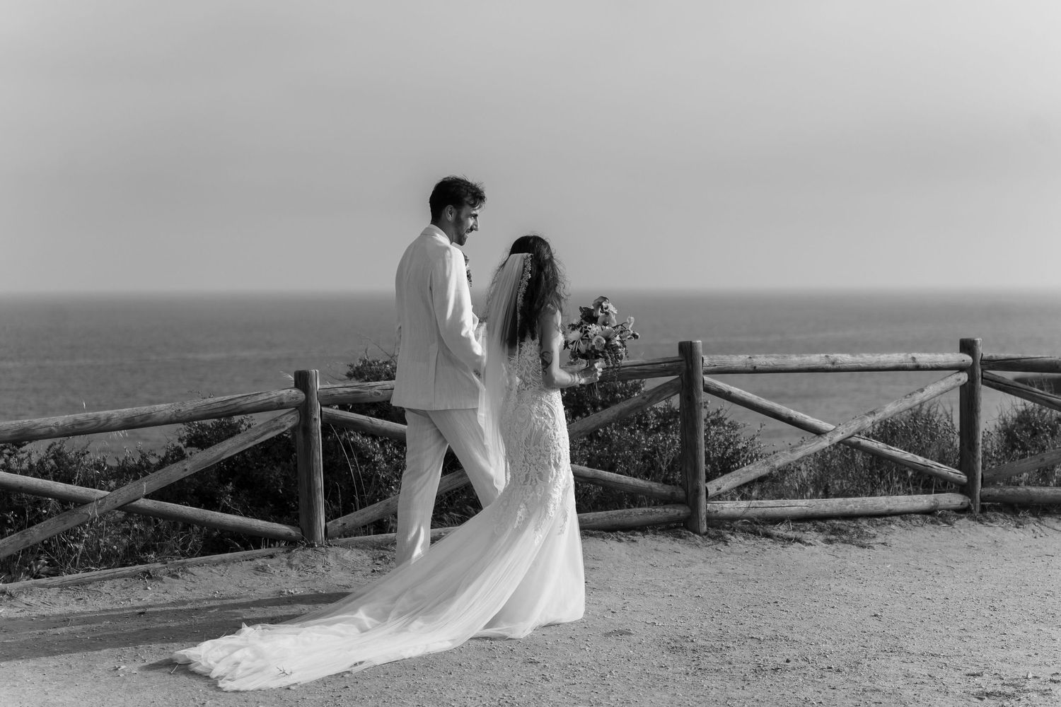 Romantic black and white portrait of bride and groom at ocean overlook with wooden fence backdrop.