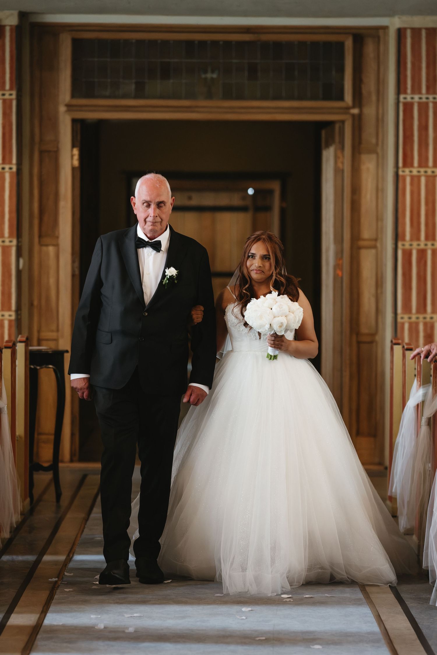 Bride in a white tulle ball gown holding a bouquet walks down the aisle with an older man in a black tuxedo.