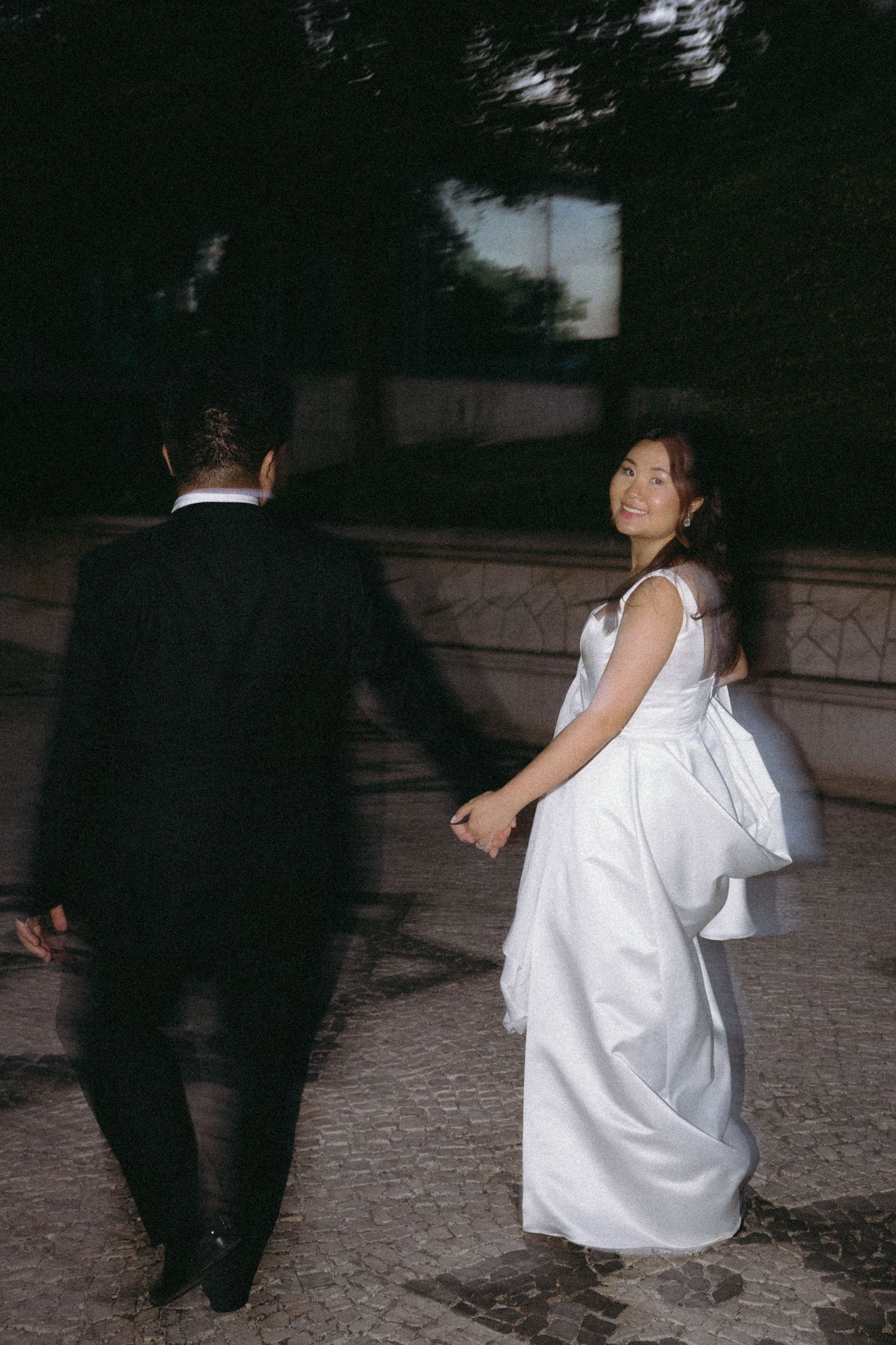 Bride in white gown with bow looks back smiling while holding groom's hand in black suit outdoors.