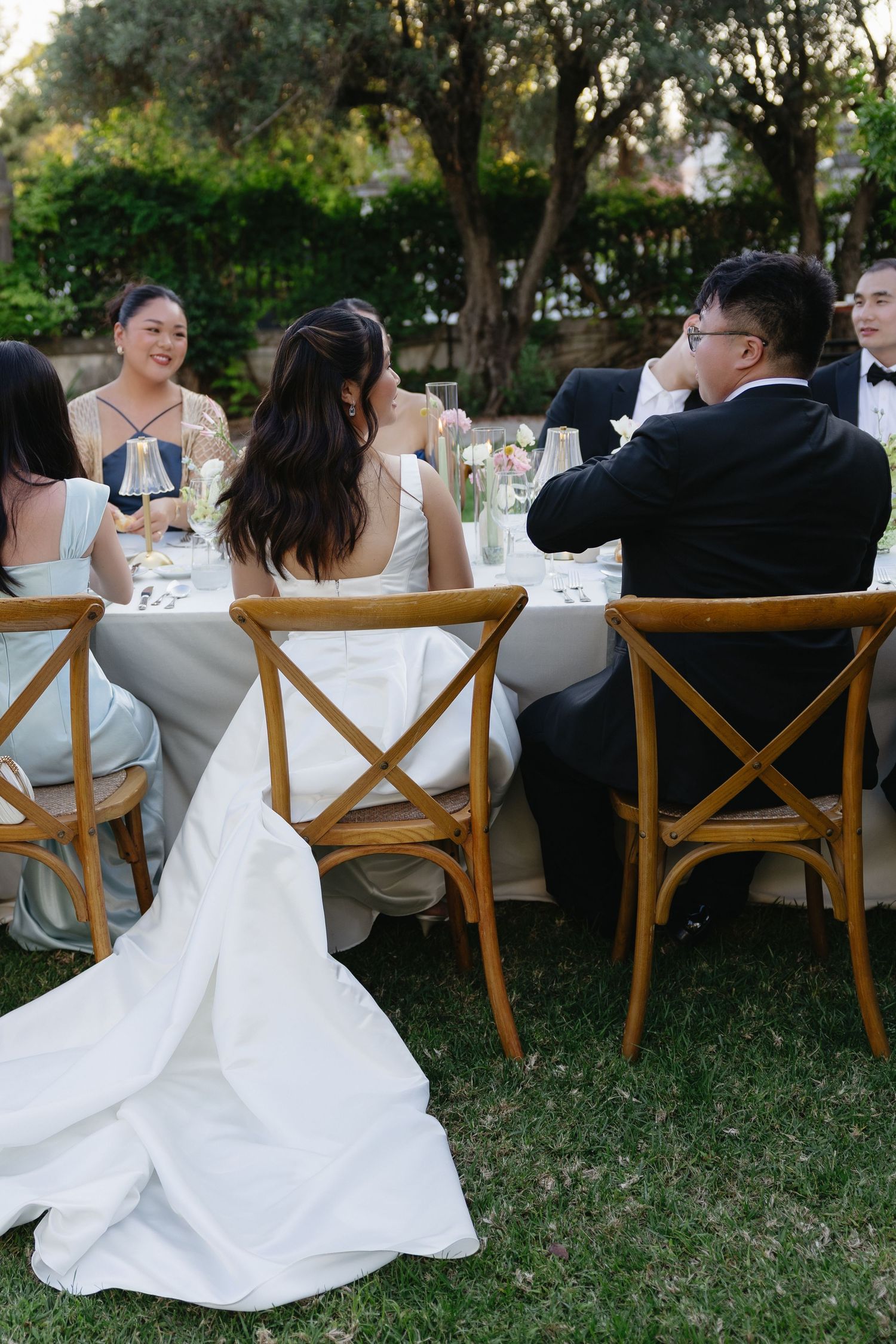 Bride in white gown and groom in tuxedo seated at outdoor wedding reception table with guests, surrounded by lush greenery.