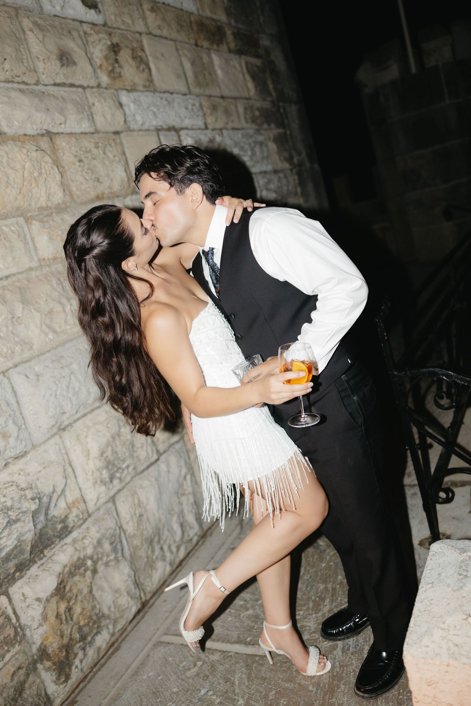 Couple sharing a romantic kiss outdoors at night, woman in white fringe dress holding champagne against stone wall.