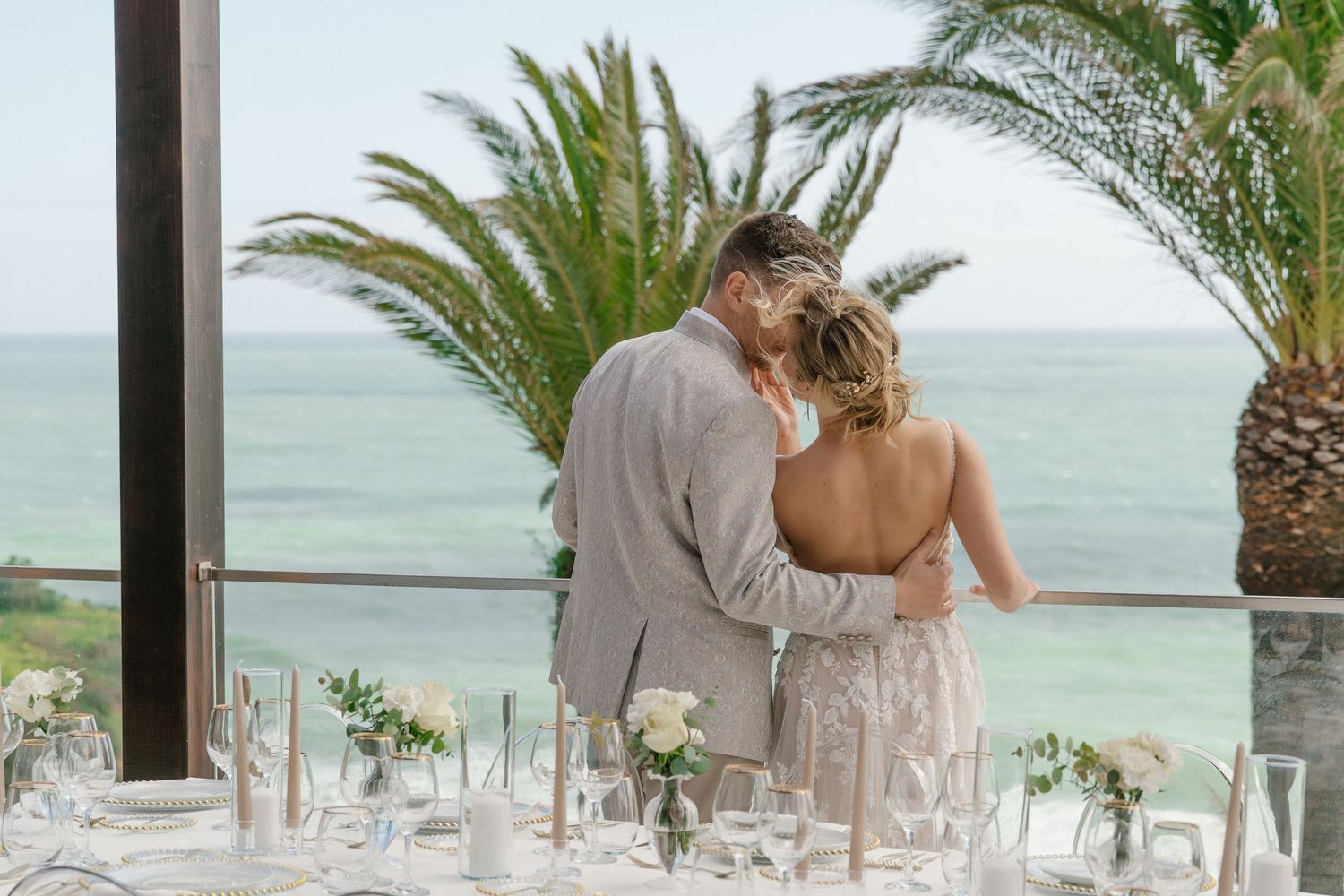 Bride and groom sharing intimate moment at beachfront reception with palm trees and ocean view backdrop.
