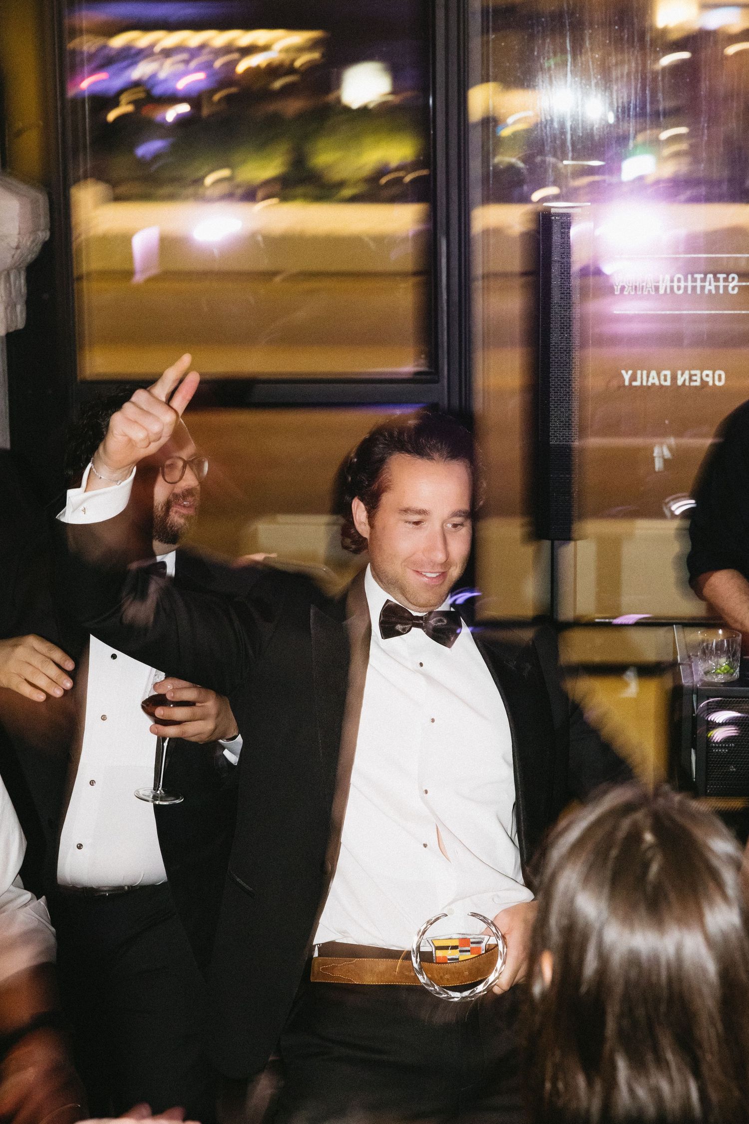 Man in tuxedo with bow tie raising hand and holding drinks at formal evening party event.