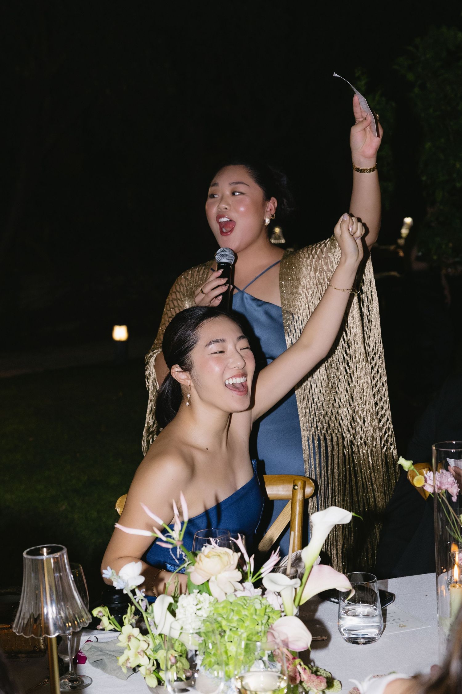 Two women in blue dresses celebrate at a wedding reception, one holding a microphone and raising her hand joyfully at night.
