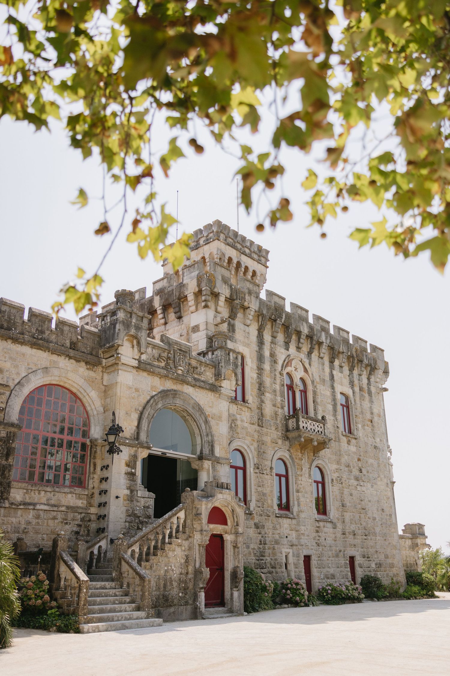 Historic stone castle with battlements, red doors, arched windows, and grand staircase entrance framed by green tree branches.