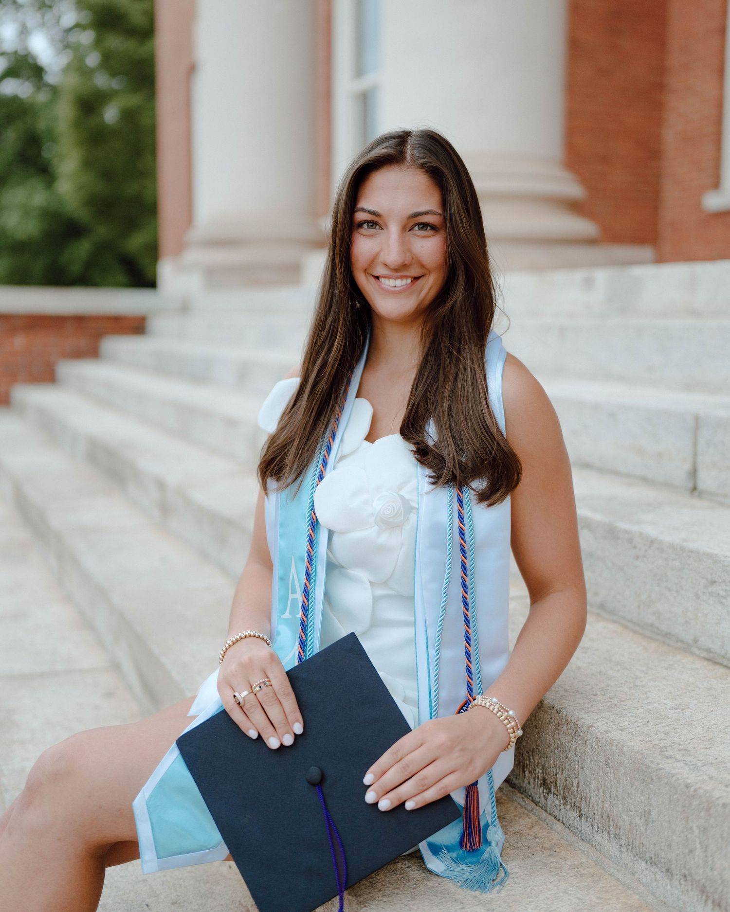 Graduate in light blue graduation gown holding diploma on stone steps of brick building with columns.
