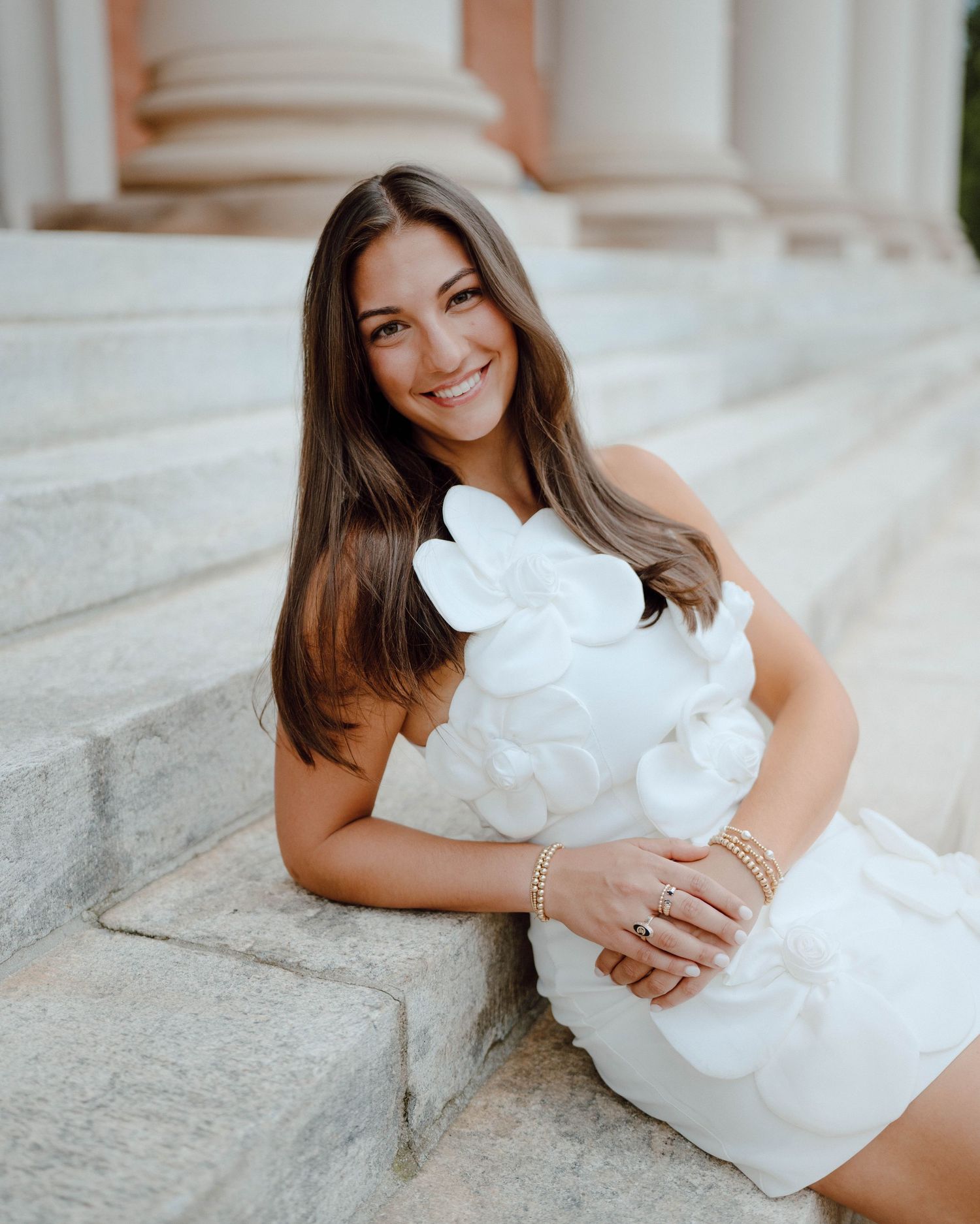 Someone in a short white dress with floral details sits on concrete steps near classical columns.