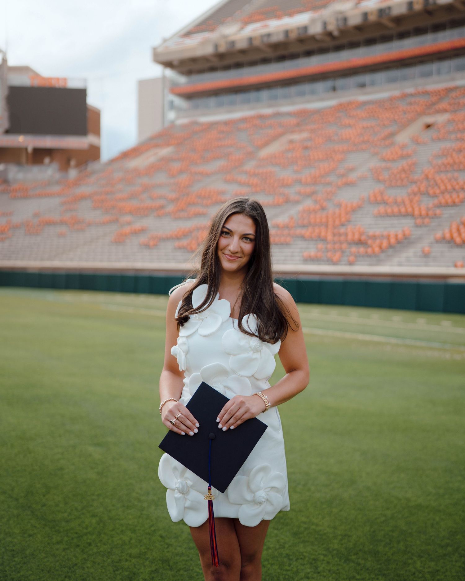 Graduate in a white dress holding a diploma on a football field with orange stadium seats in the background.