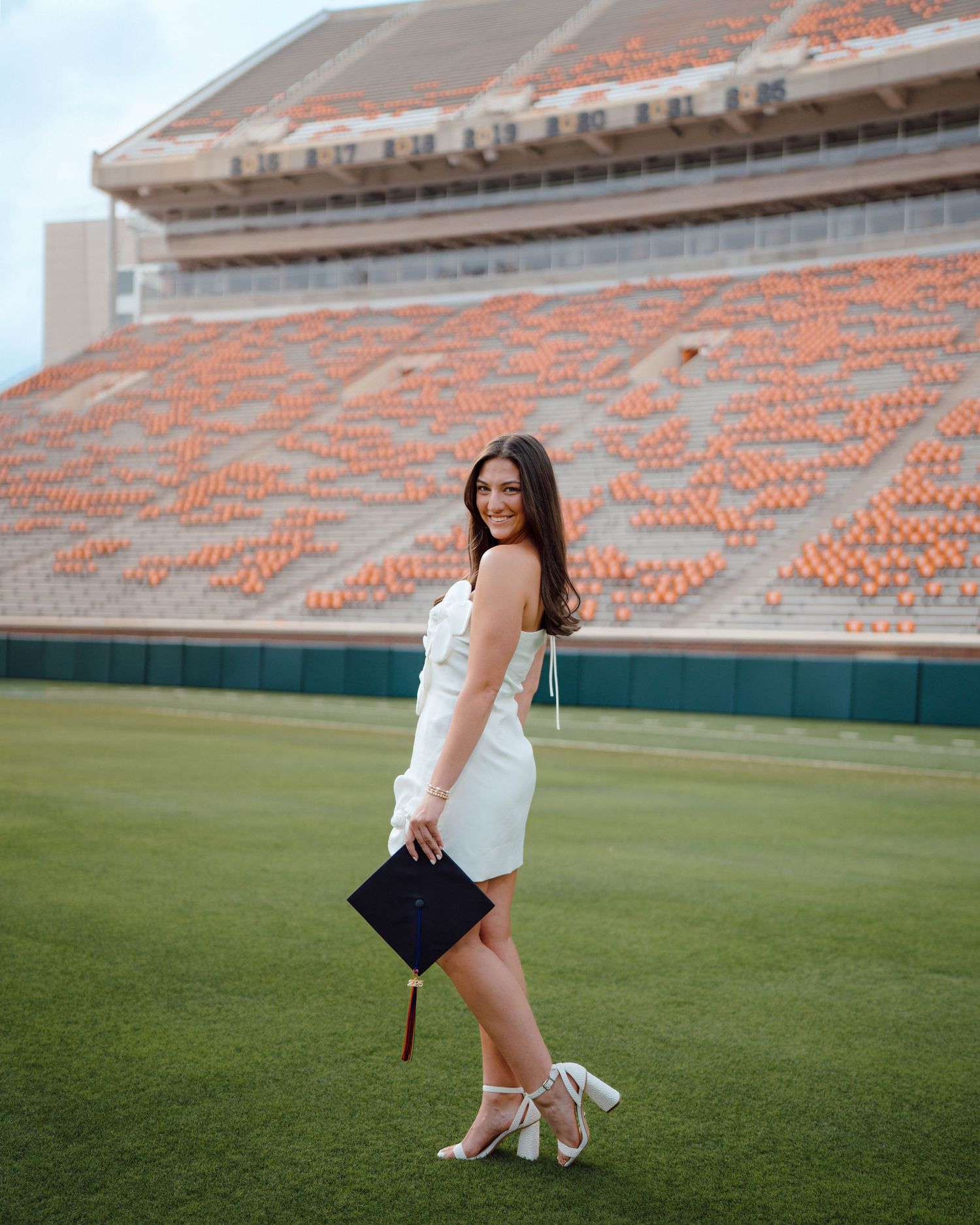 Graduate poses in white dress holding diploma on bright green football field with stadium seats in background.