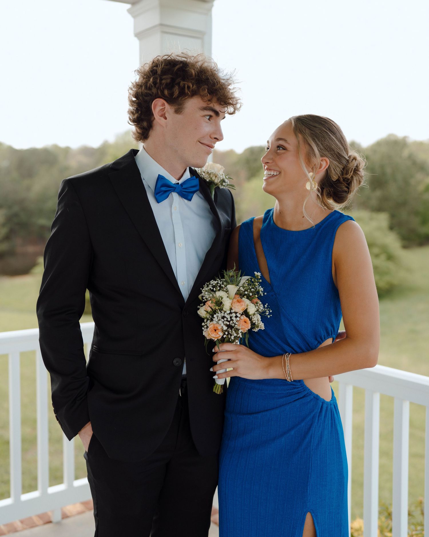 A young couple in formal attire share a sweet moment on a white porch, with her in royal blue and him in a bow tie.