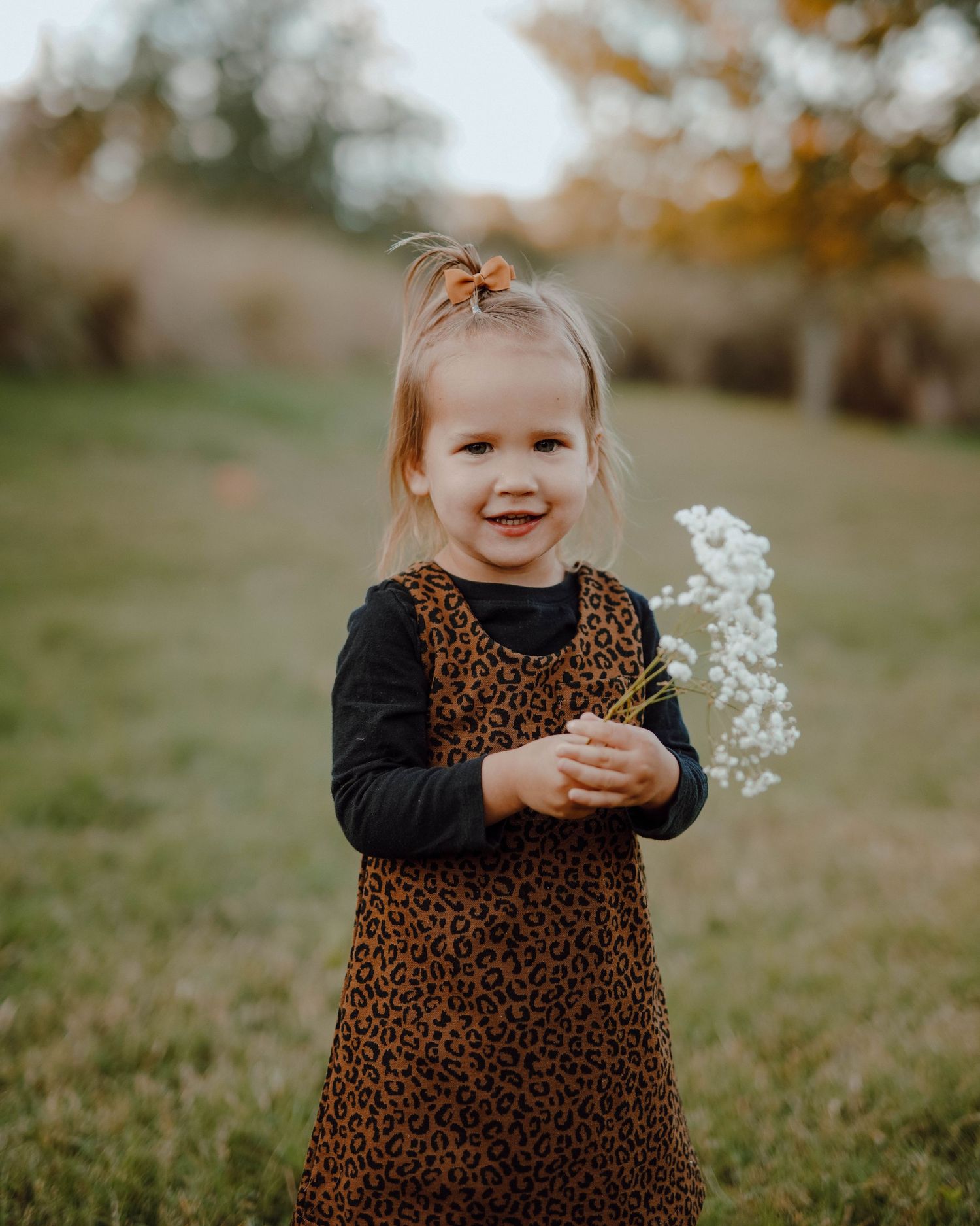 A little one wearing a leopard print dress holds white flowers while standing in a grass field during golden hour.