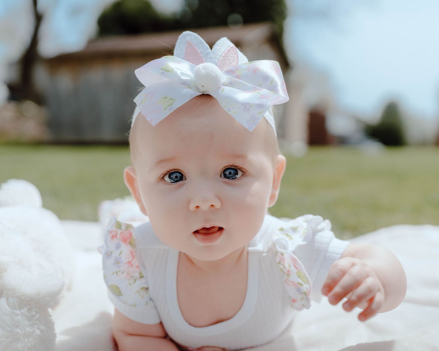 A baby wearing a white lace dress and bow lies on a white blanket outdoors on a sunny day.