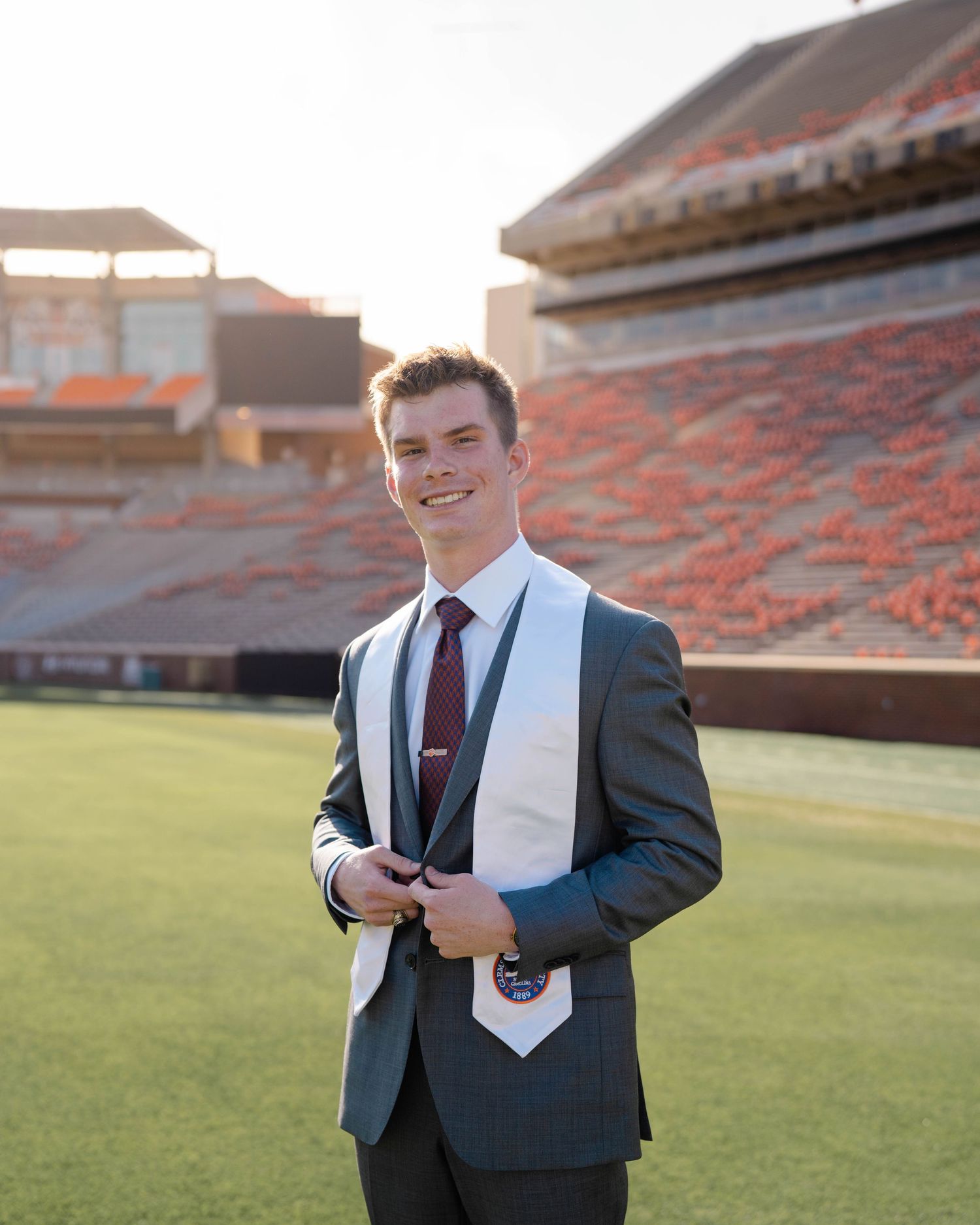Professional graduate portrait in suit and tie standing on football field with stadium seats in background at sunset.