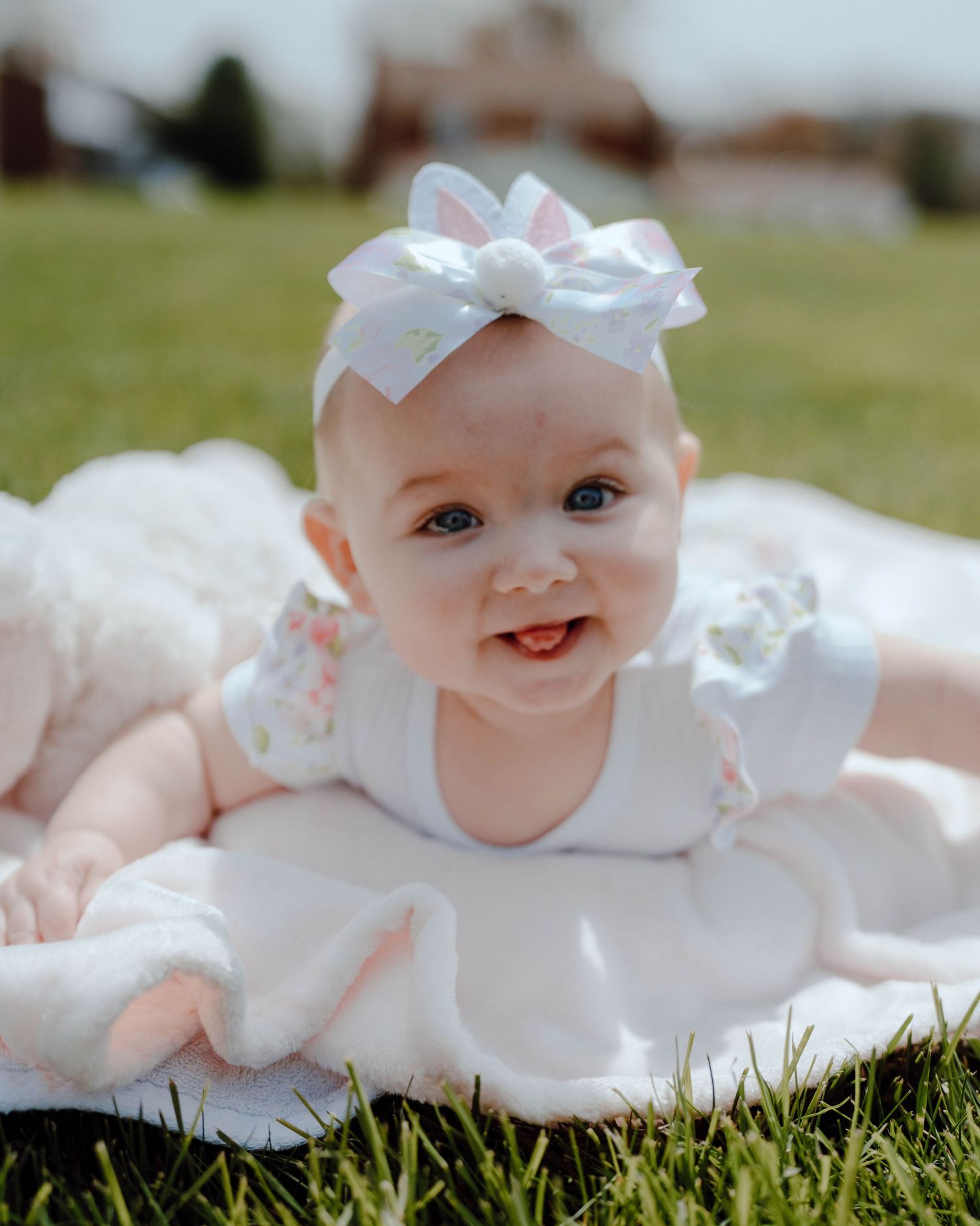 Smiling baby wearing a white dress and bow headband lies on grass during an outdoor photo session.