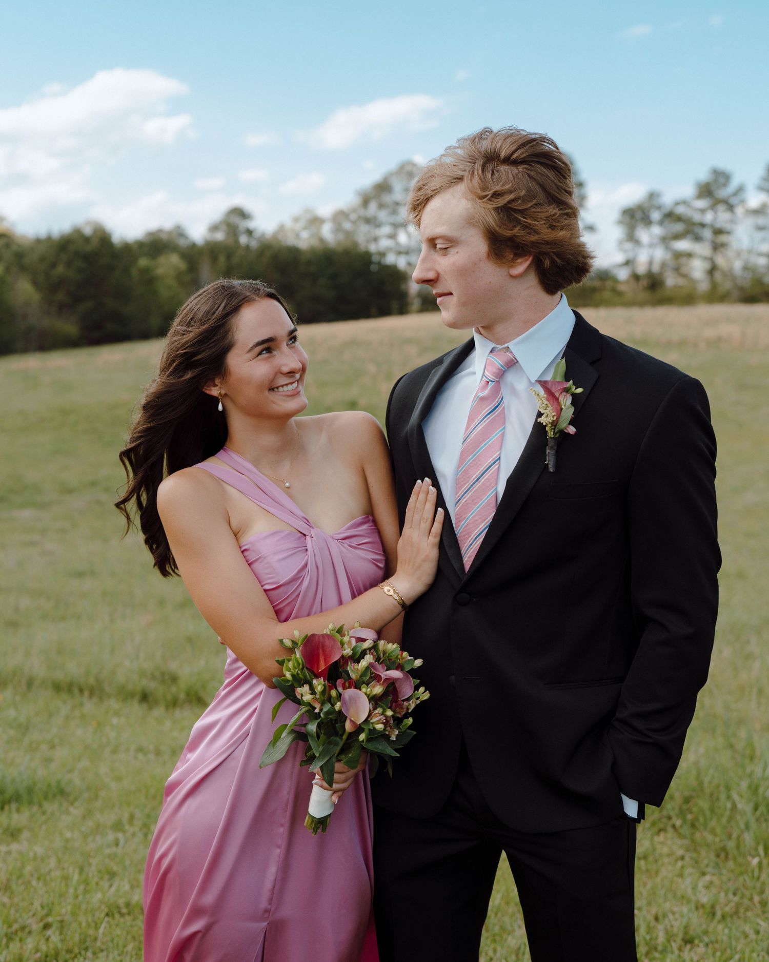 A couple in formal attire stands in a grassy field, with her holding a pink bouquet and wearing a pink dress.