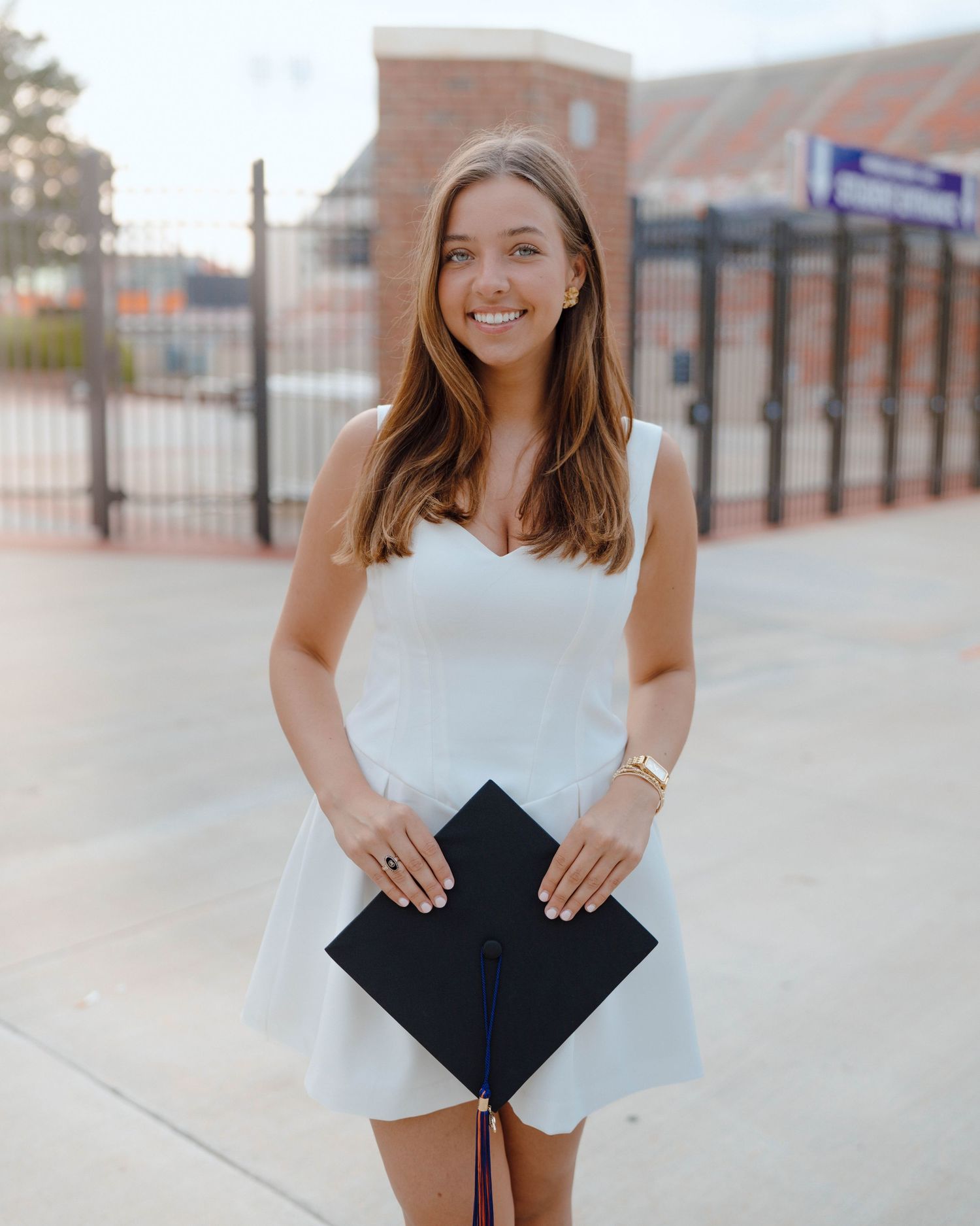 Graduate in white dress stands with graduation cap against campus building backdrop.