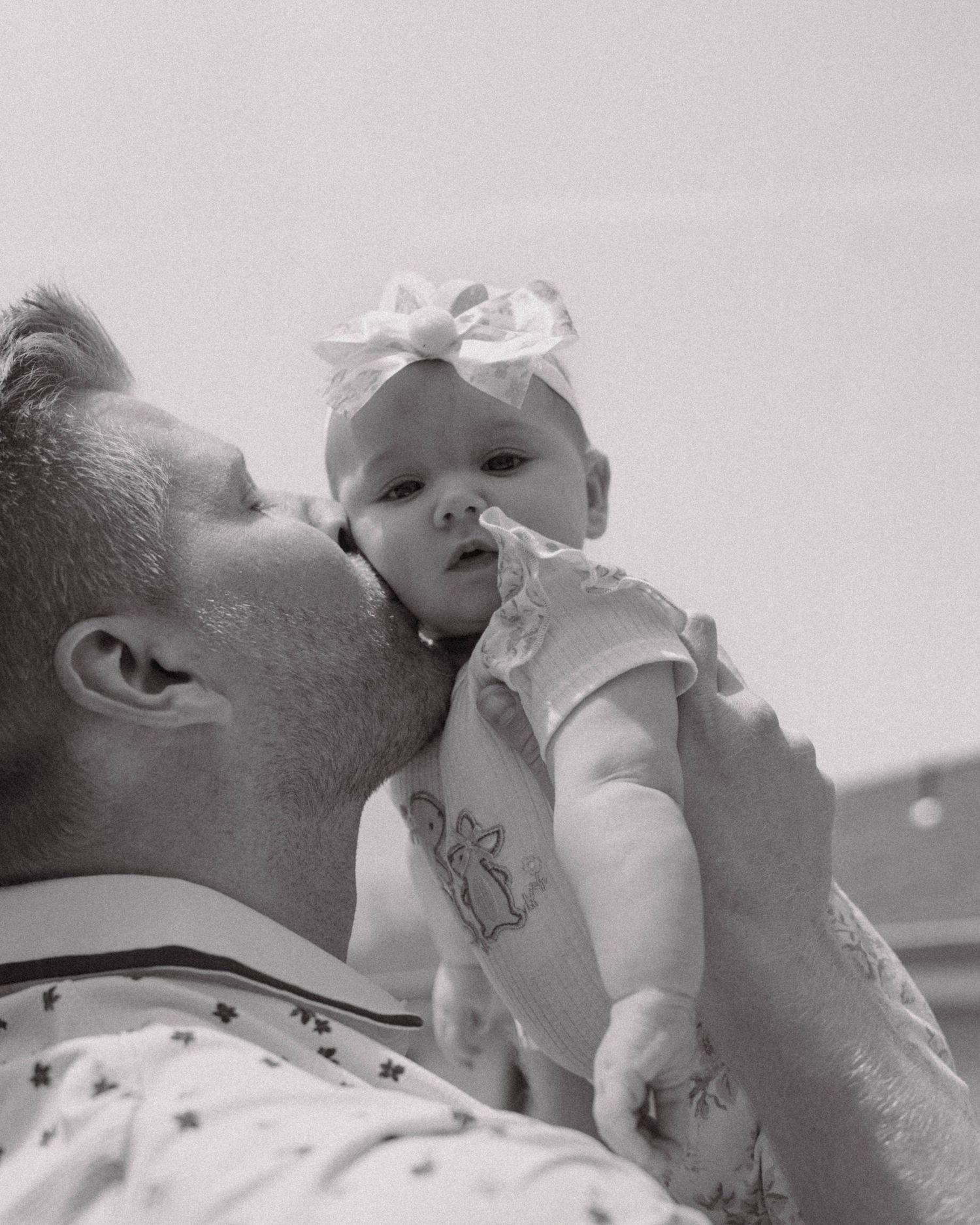 A tender black and white photo shows a tattooed parent gently kissing their baby wearing a bow in an intimate moment.