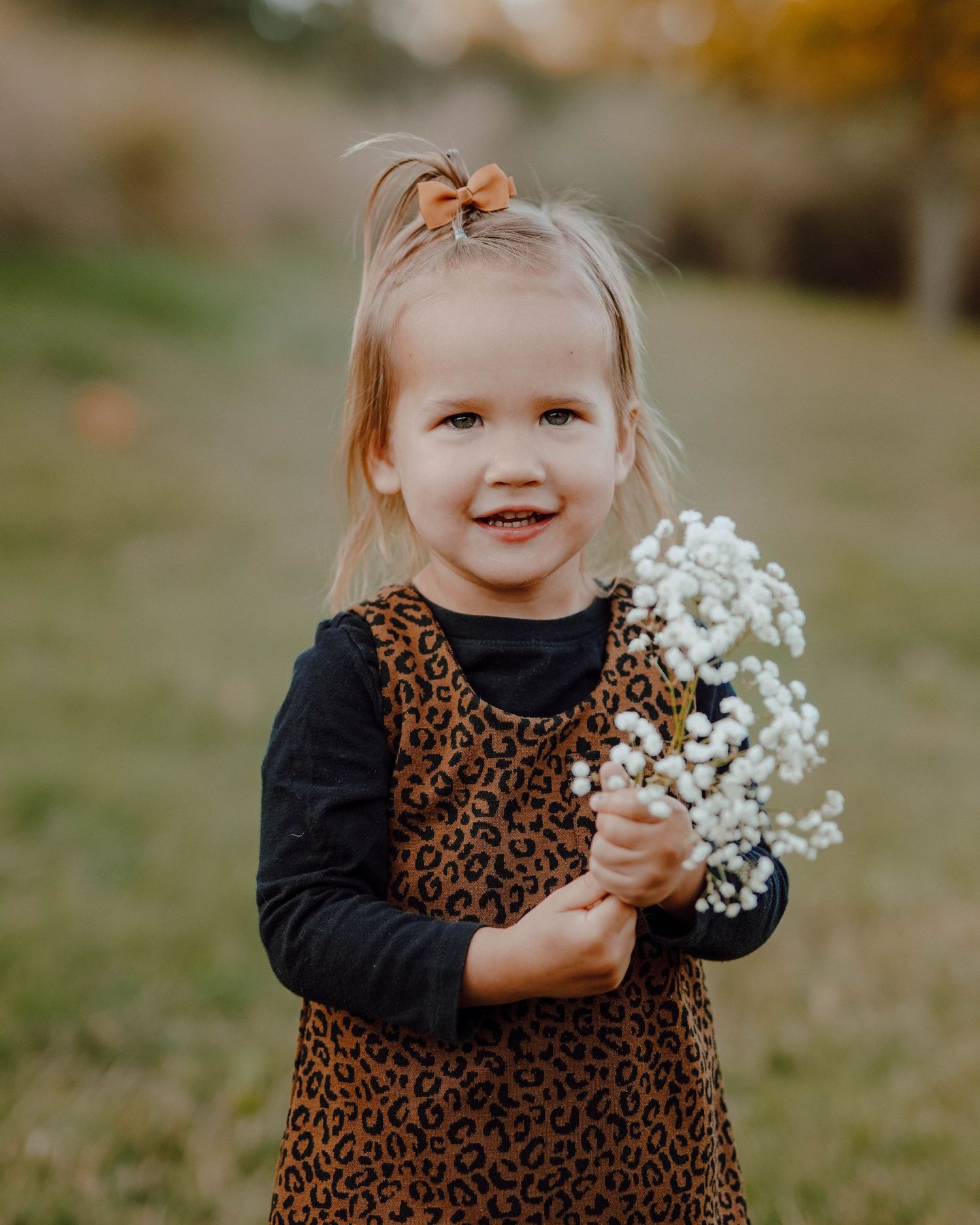 Small child in leopard print dress and black shirt holds delicate white baby's breath flowers while standing outdoors.