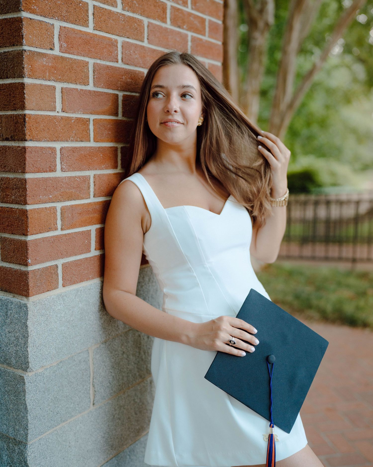 Graduate in white dress poses with diploma holder against brick wall on sunny day outdoors.