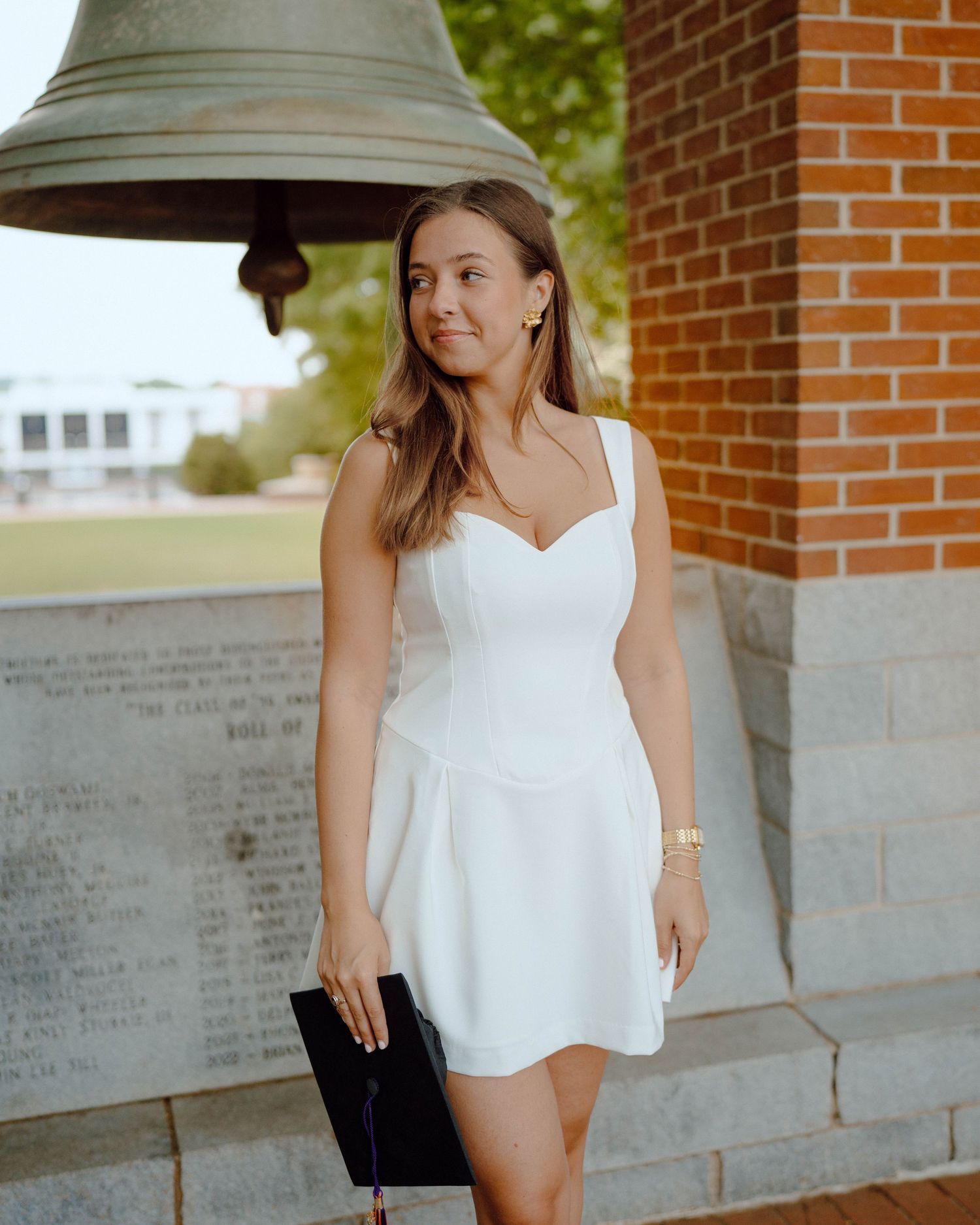 A person in an elegant white summer dress poses next to a large bell in front of a brick building on a sunny day.