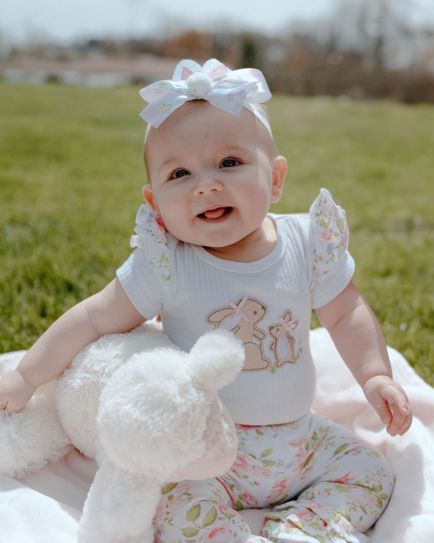 Smiling baby in a white bow and floral outfit sits outdoors with a plush bunny toy on a blanket.