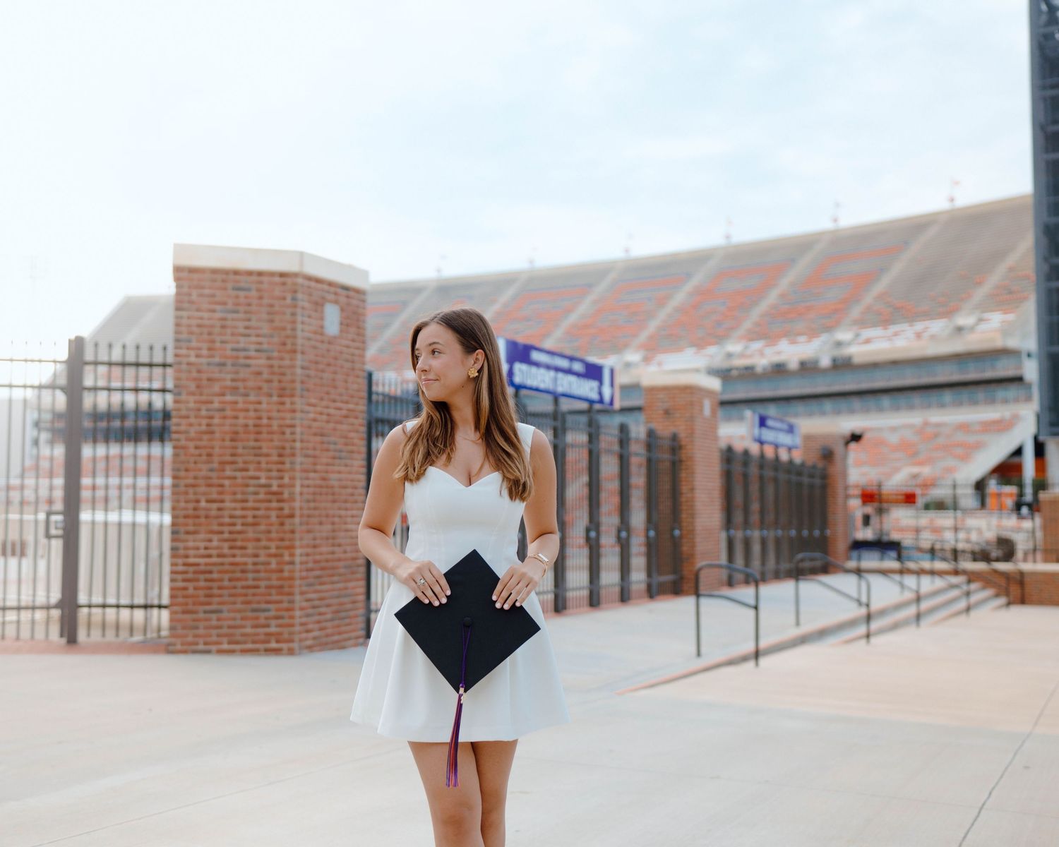 Standing in front of a brick stadium entrance in a white dress holding a graduation diploma cover on a sunny day.