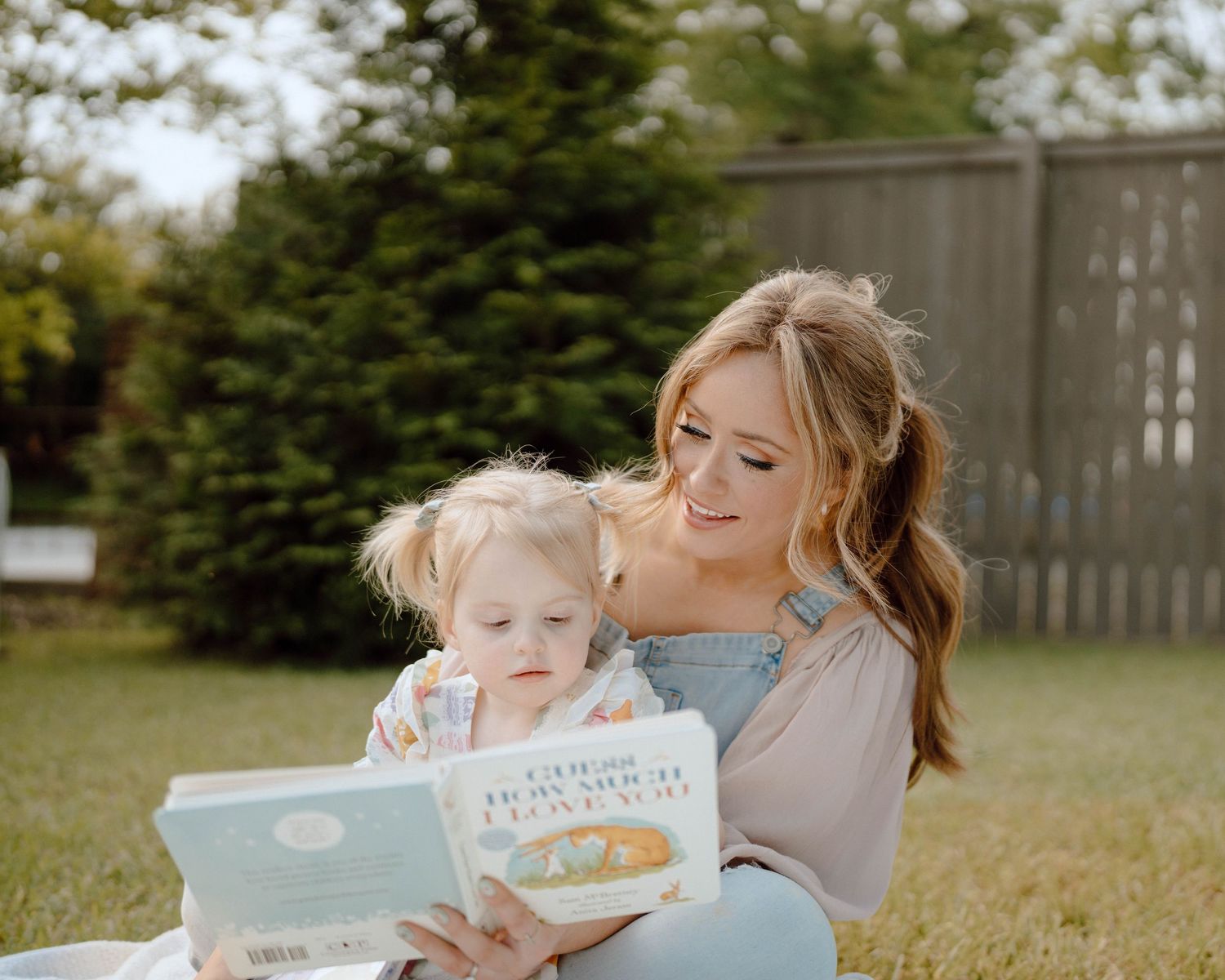 A parent and child sit in a grassy backyard while reading a children's picture book together on a sunny day.