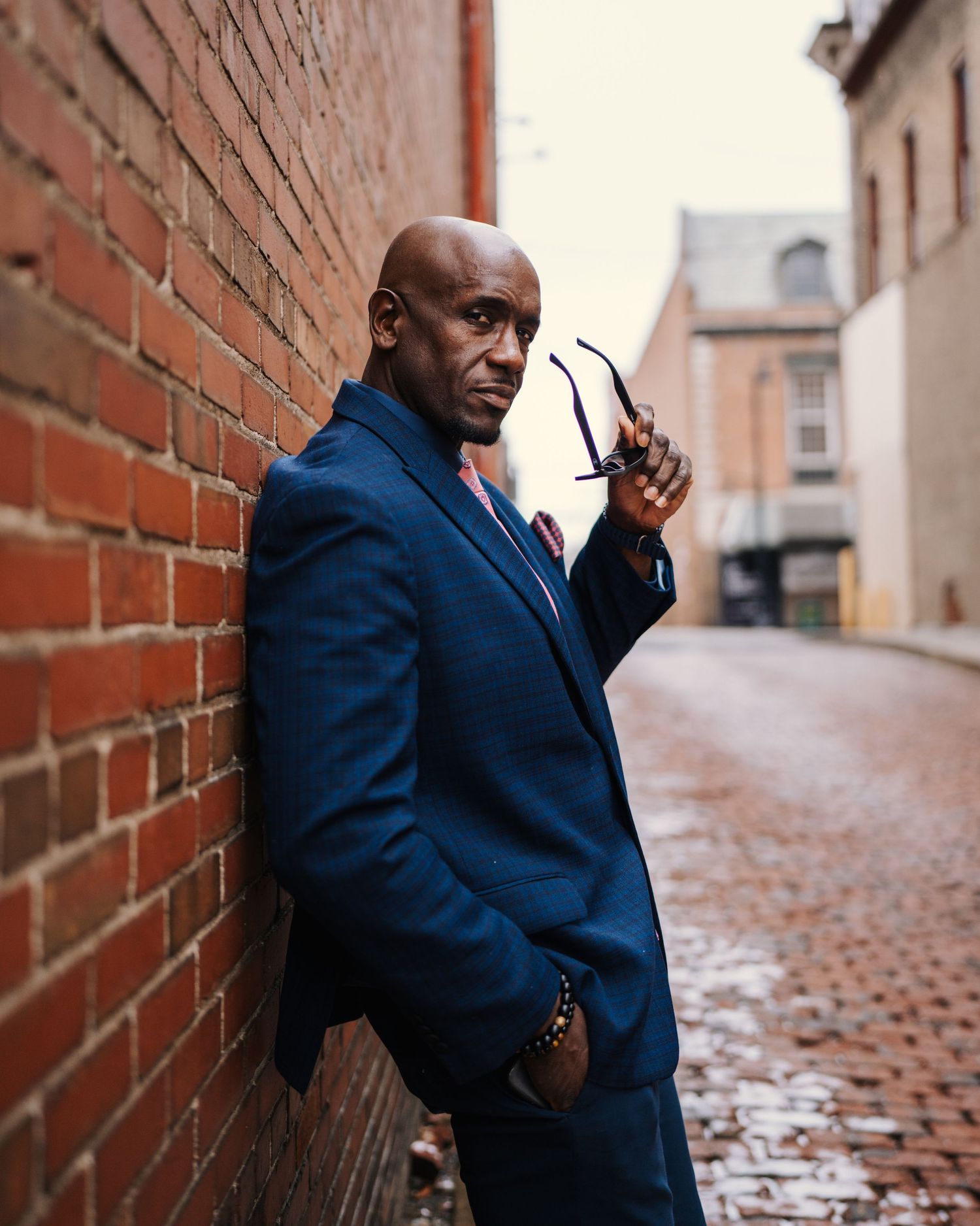 Stylish businessman in navy suit leaning against brick wall holding glasses.