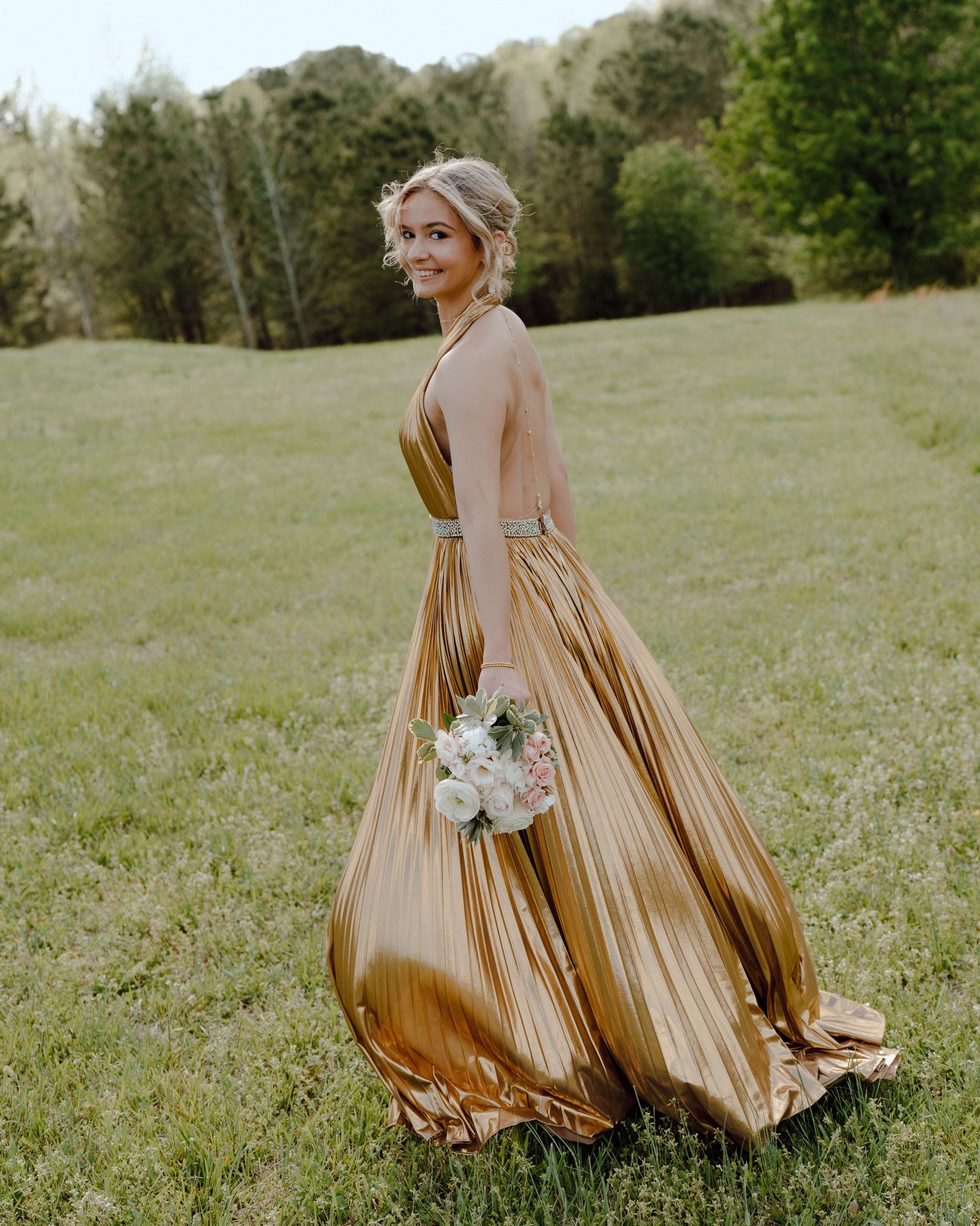 Someone wearing a flowing champagne gold formal gown poses with a white bouquet in a grassy field at sunset.